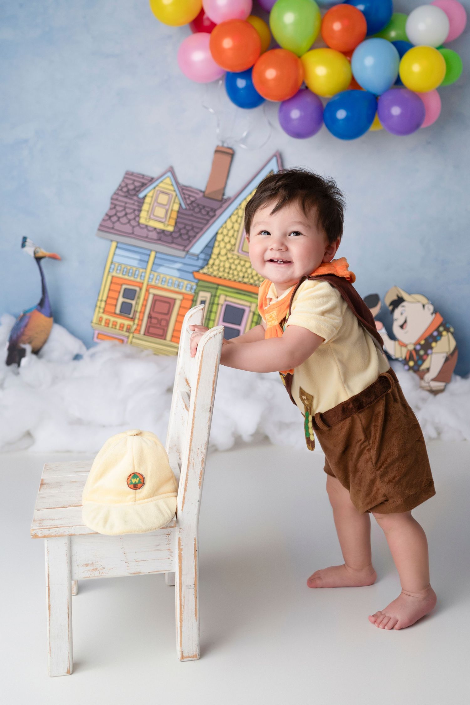 Whimsical photo setup with colorful balloons, cartoon houses, and a distressed white chair against cloudy backdrop.