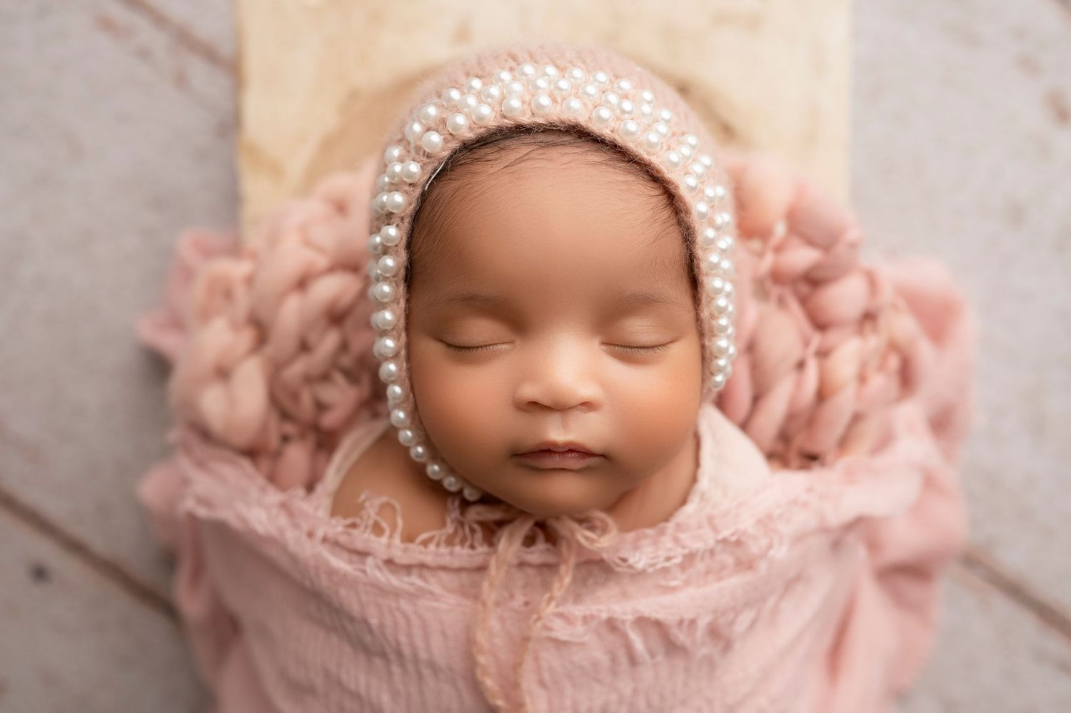 Sleeping newborn baby wrapped in soft pink blanket wearing delicate pearl headband during professional photoshoot.