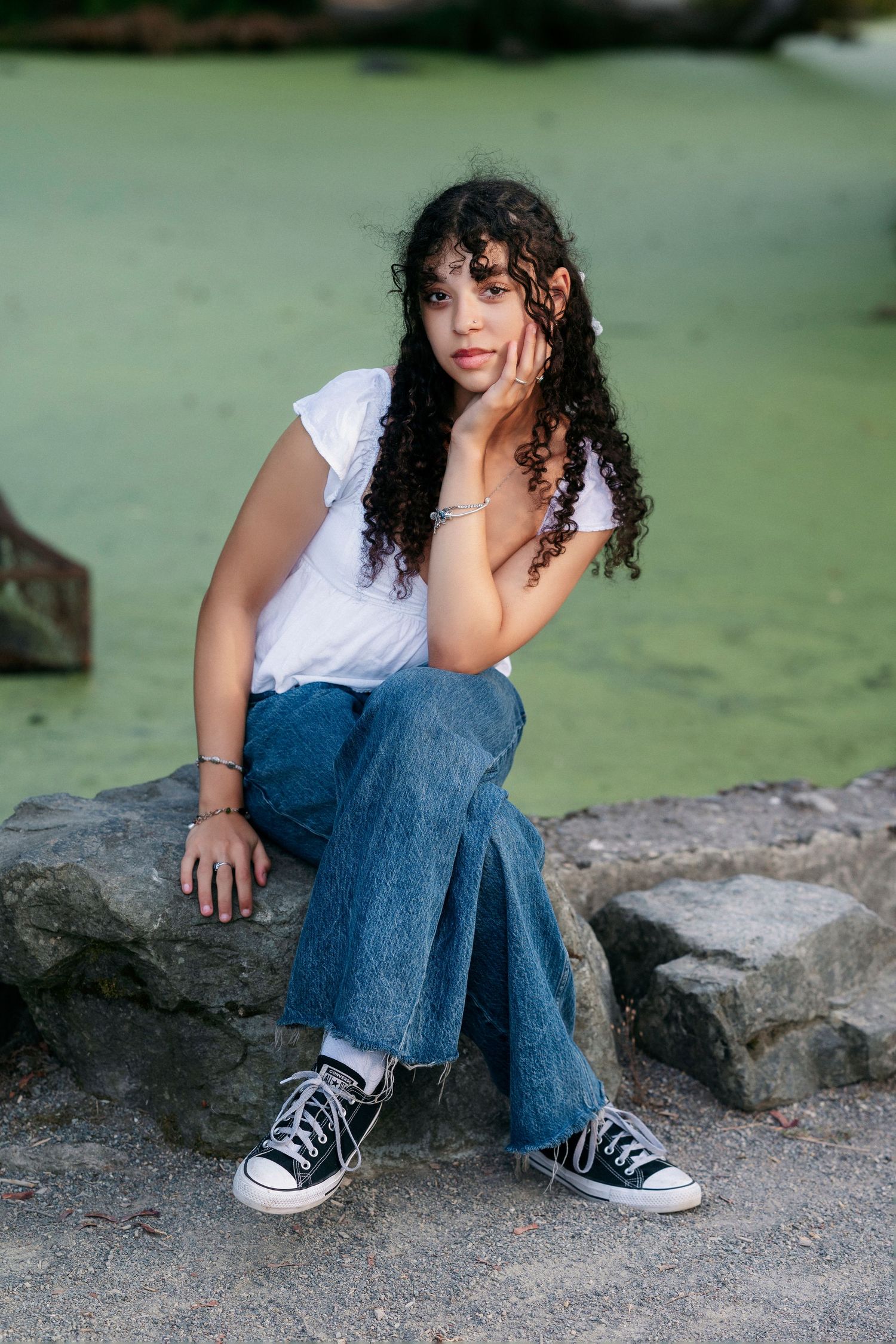A person in a white top and jeans sits casually on rocks near a tranquil pond.