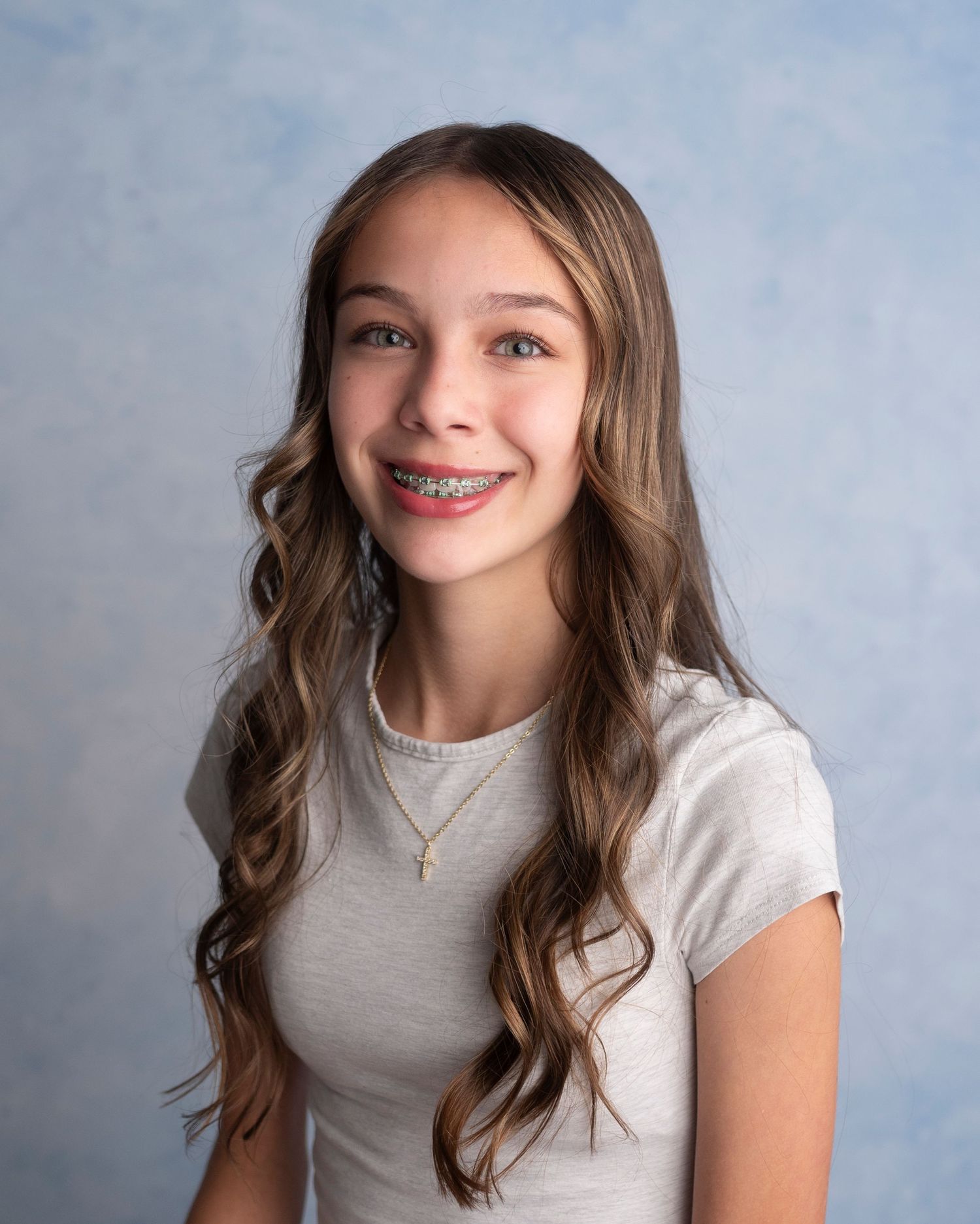Portrait of a young person with long wavy brown hair wearing a white t-shirt and smiling against a blue background.