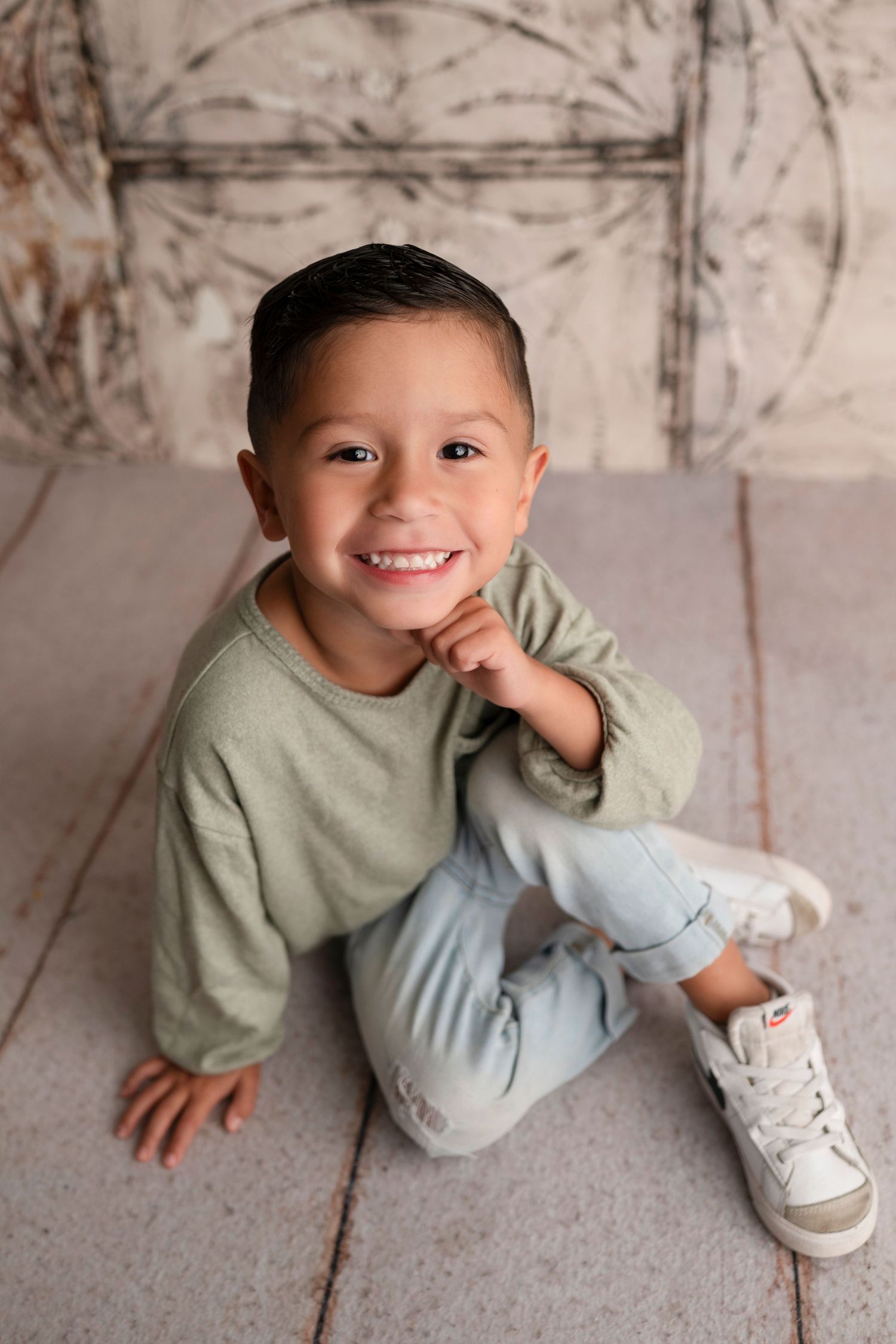 A child in a sage green sweater and light blue pants lies playfully on a textured gray floor.