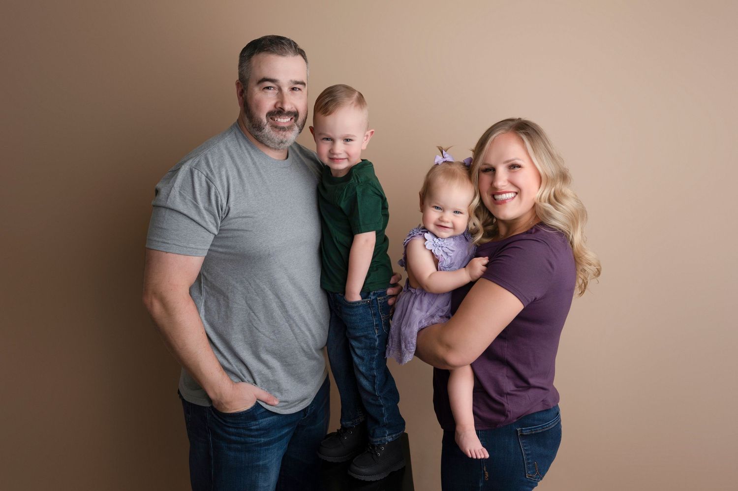 Happy family of four standing together, dad and mom with toddler son and baby daughter in lavender outfit.