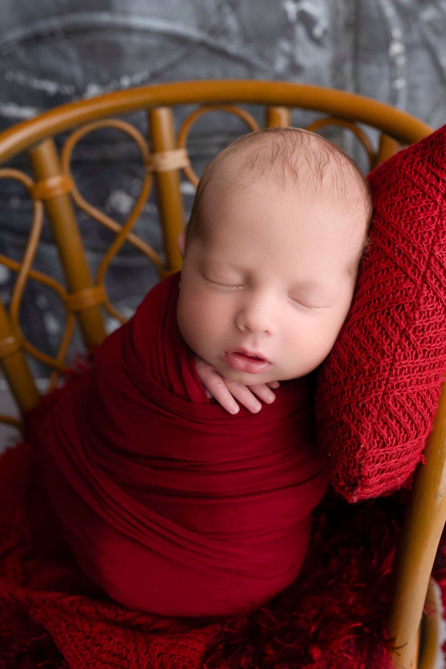 Newborn baby swaddled in red wrap sleeping peacefully in a small rattan chair with a red pillow.