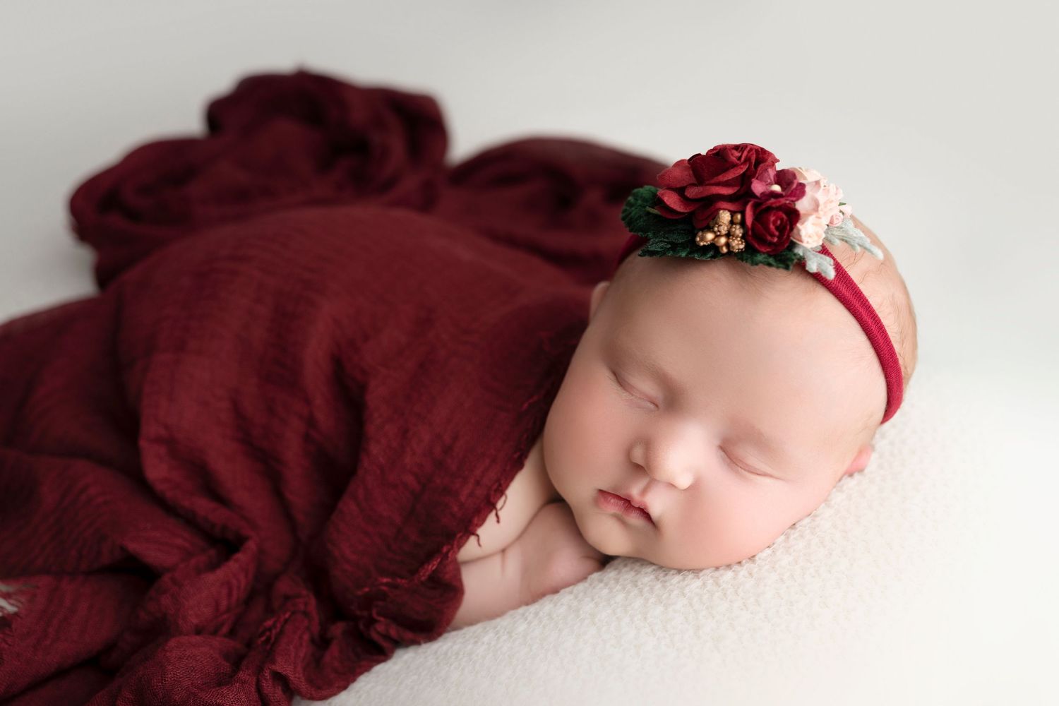 A sleeping newborn in burgundy fabric with a small red bow headband rests peacefully on white bedding.