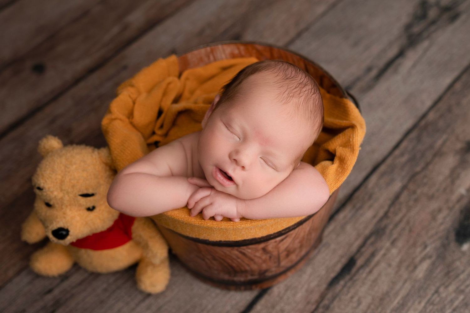 Sleeping newborn baby curled in a wooden bucket with orange fabric and a Winnie the Pooh teddy bear.