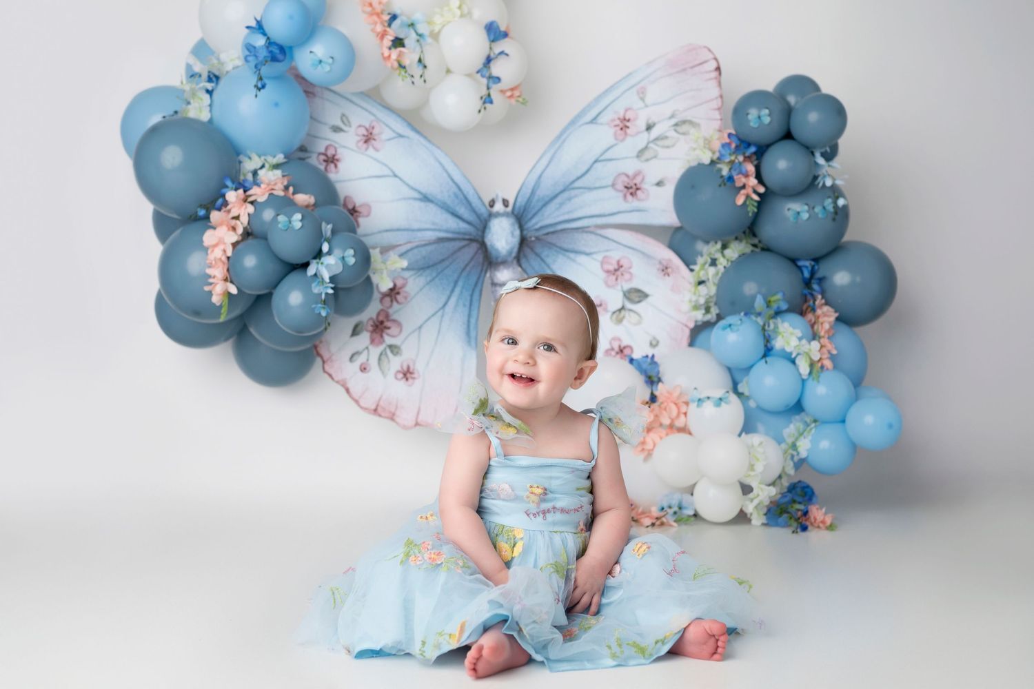 Decorative backdrop with blue balloons, flowers, and butterfly accents creates a dreamy scene for photography.