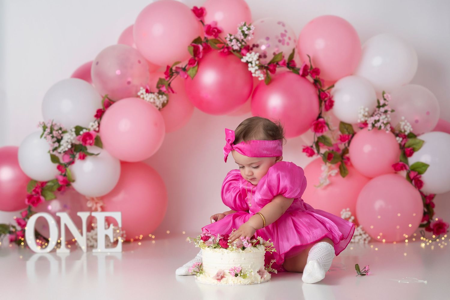 Pink and white balloon garland with floral accents and glittery ONE text decoration creates a festive party backdrop.