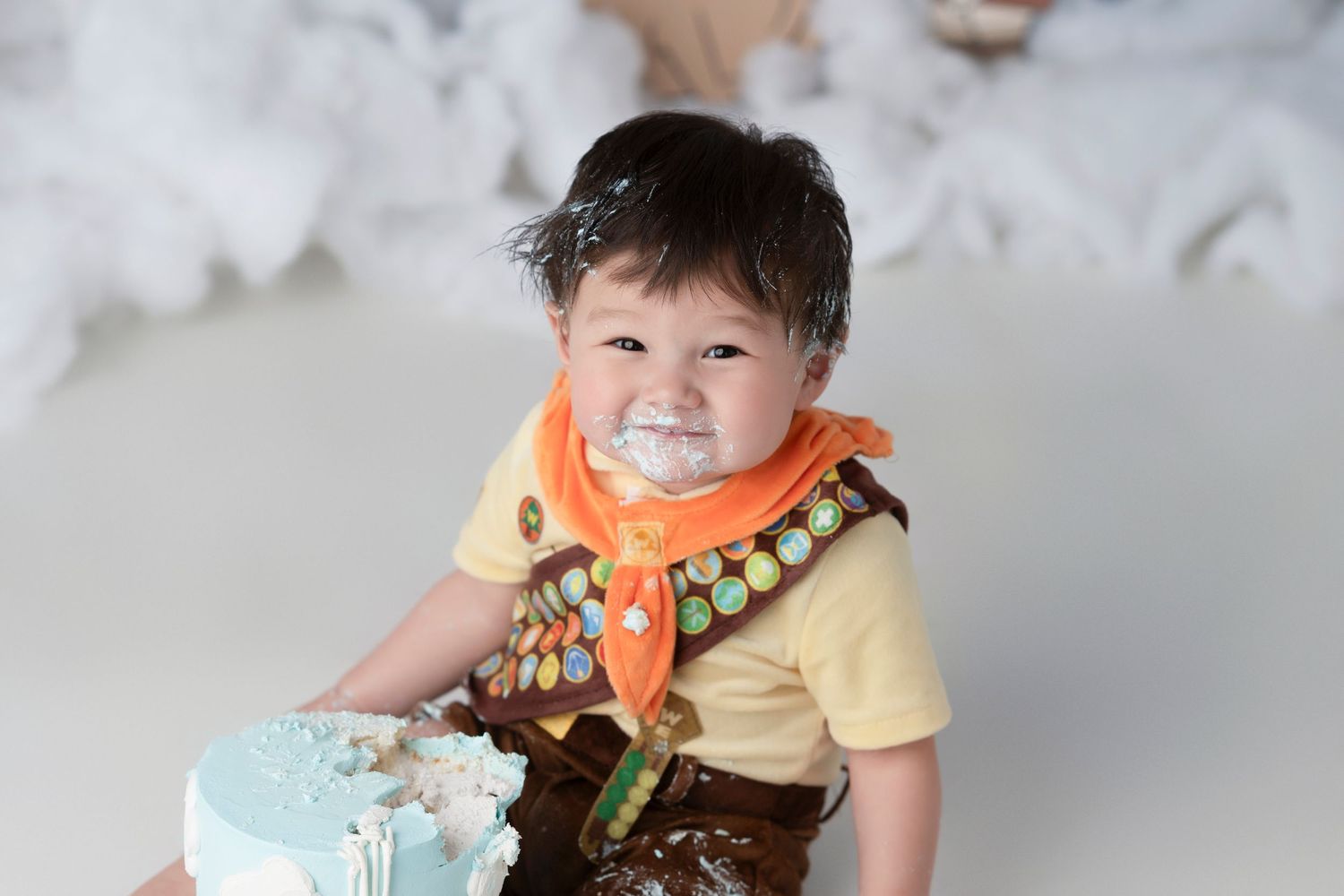 A baby wearing a brown and orange outfit smiles while sitting with a blue birthday cake.