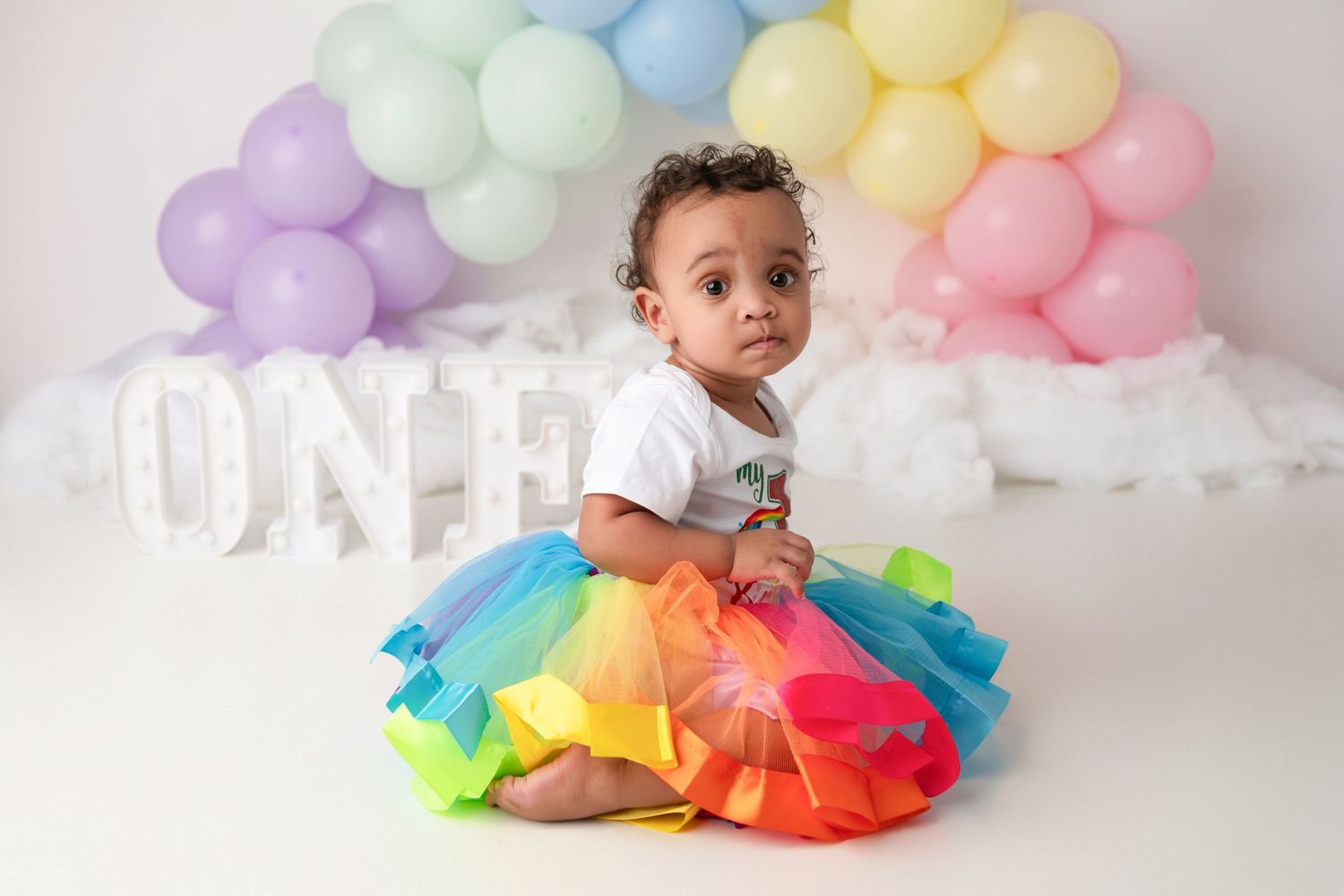 Rainbow tulle tutu dress and pastel balloon clouds create a whimsical first birthday photoshoot setup.