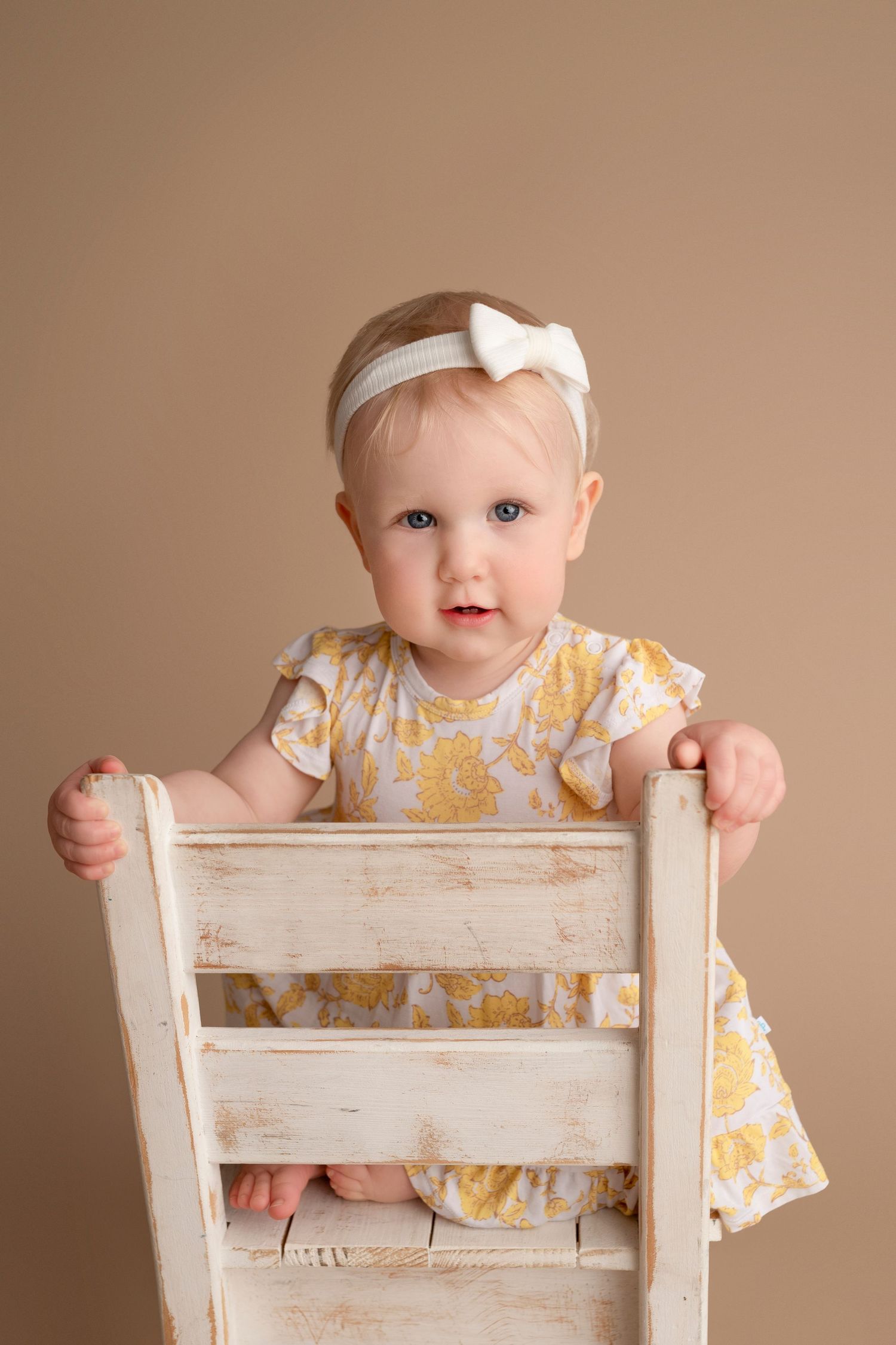 Baby in a yellow floral dress sits in a distressed white wooden chair against a neutral backdrop.