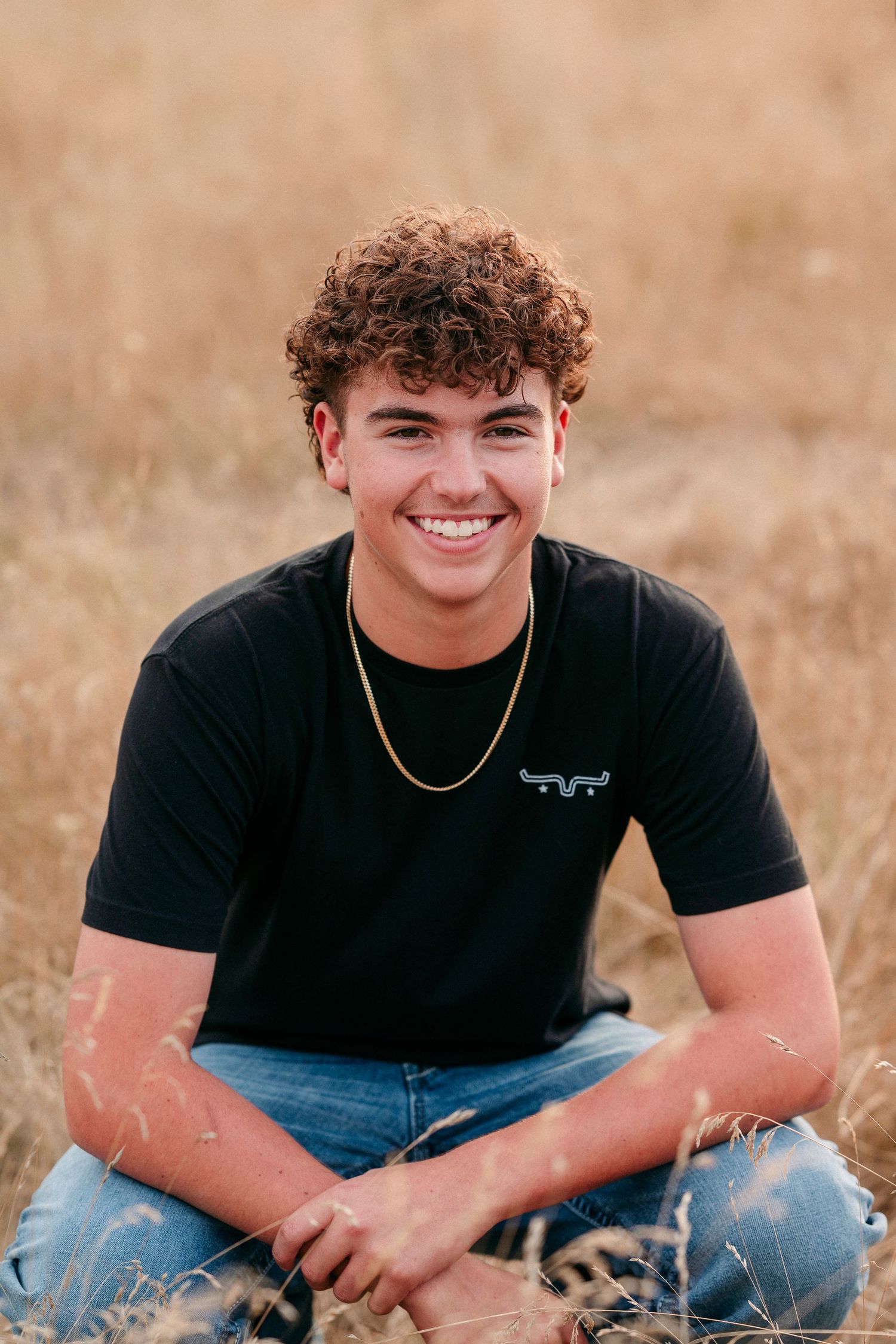 A person wearing a black t-shirt and jeans sits casually in a grassy outdoor setting during golden hour.