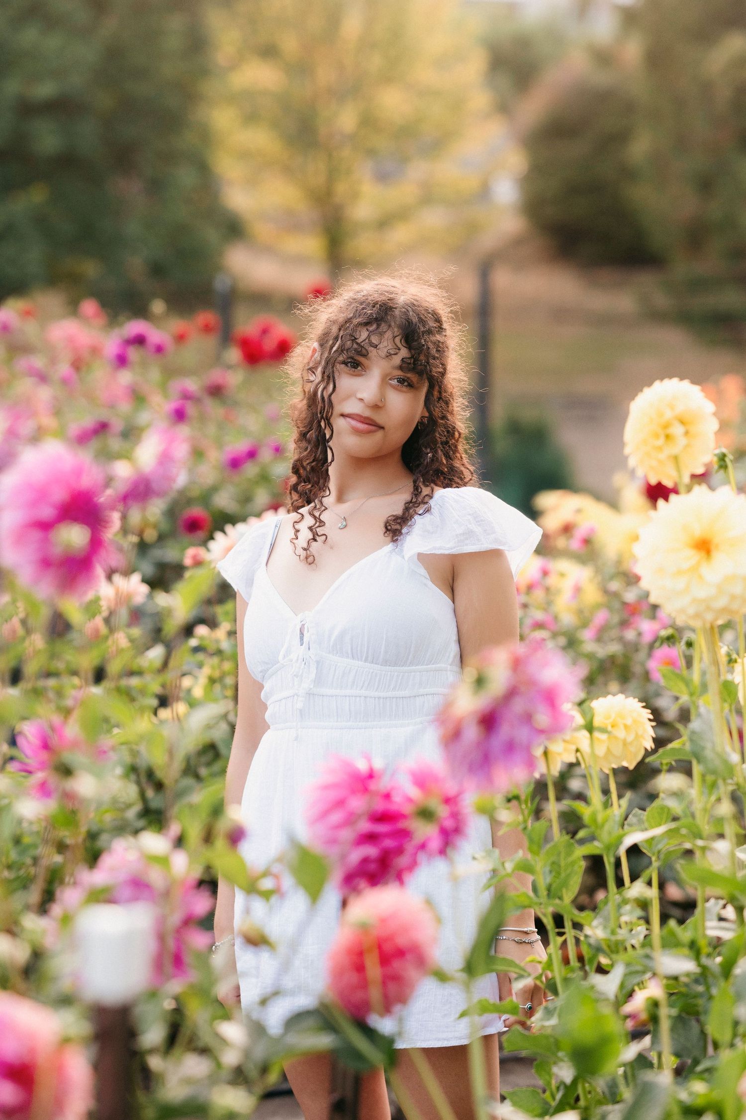 A person in a white summer dress poses among vibrant pink and yellow flowers in a garden setting.