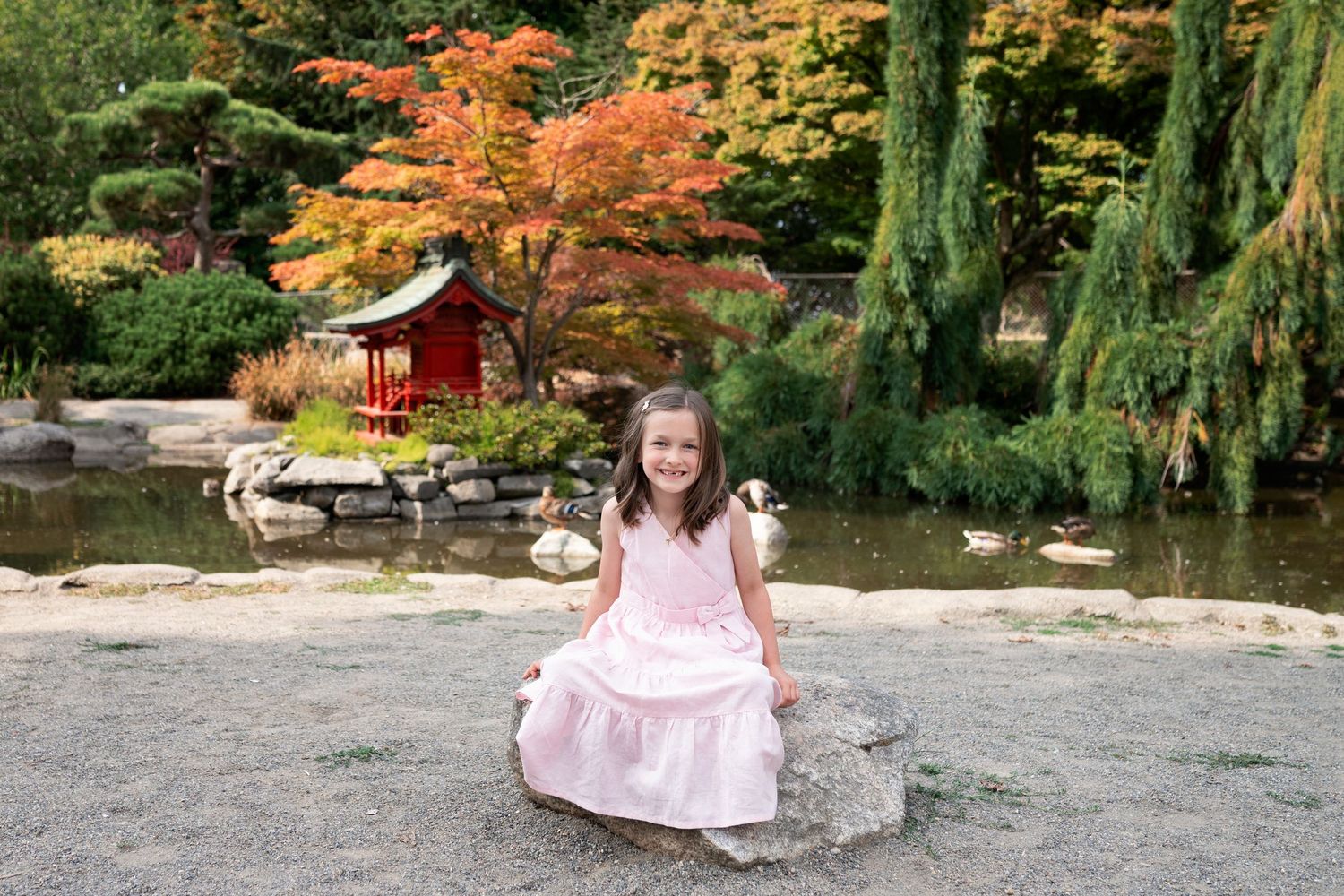 Someone in a pink dress poses in a tranquil garden setting with autumn foliage and stone pathways.
