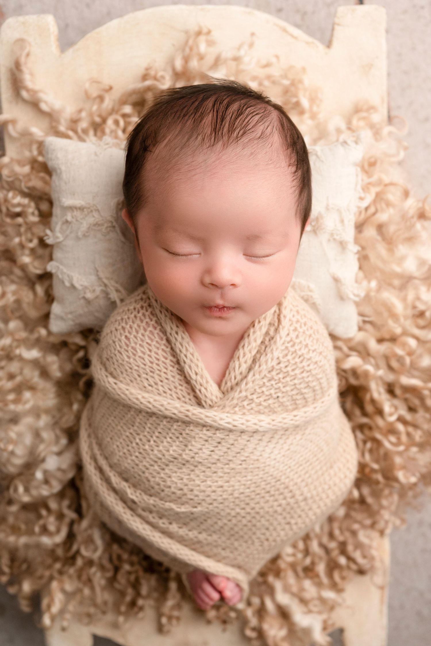 Sleeping newborn wrapped in cozy beige knit blanket on cream colored textured backdrop.