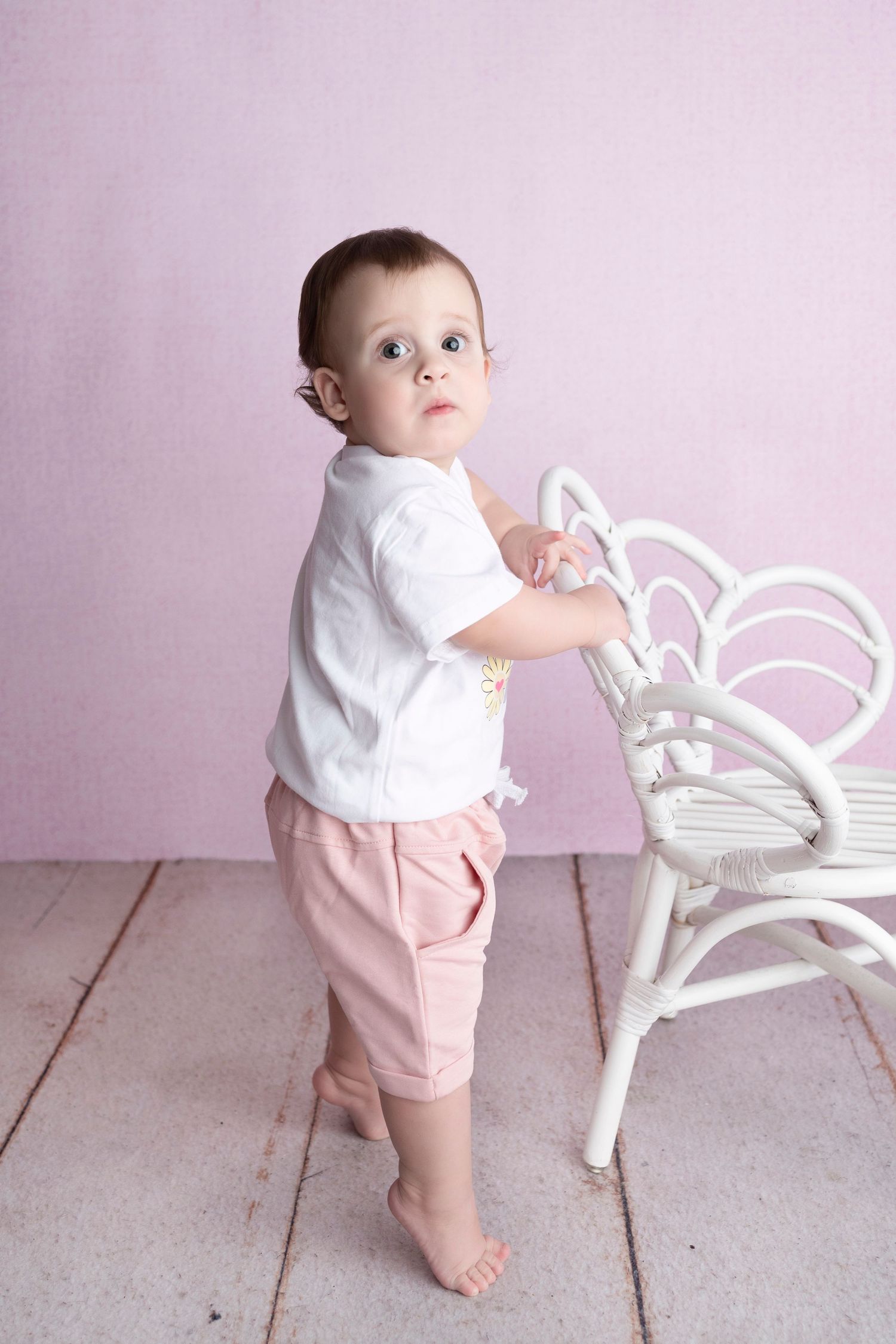 Baby in white shirt and pink pants stands barefoot next to white wicker chair against pink wall.