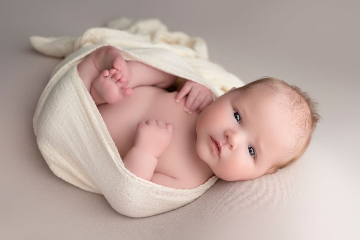 Newborn baby wrapped in a soft white swaddle blanket, lying on a gray background with blue eyes open.
