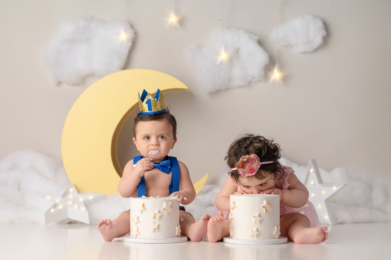 Two babies in party hats sit beside small white cakes with a moon and stars backdrop for a first birthday smash cake session.