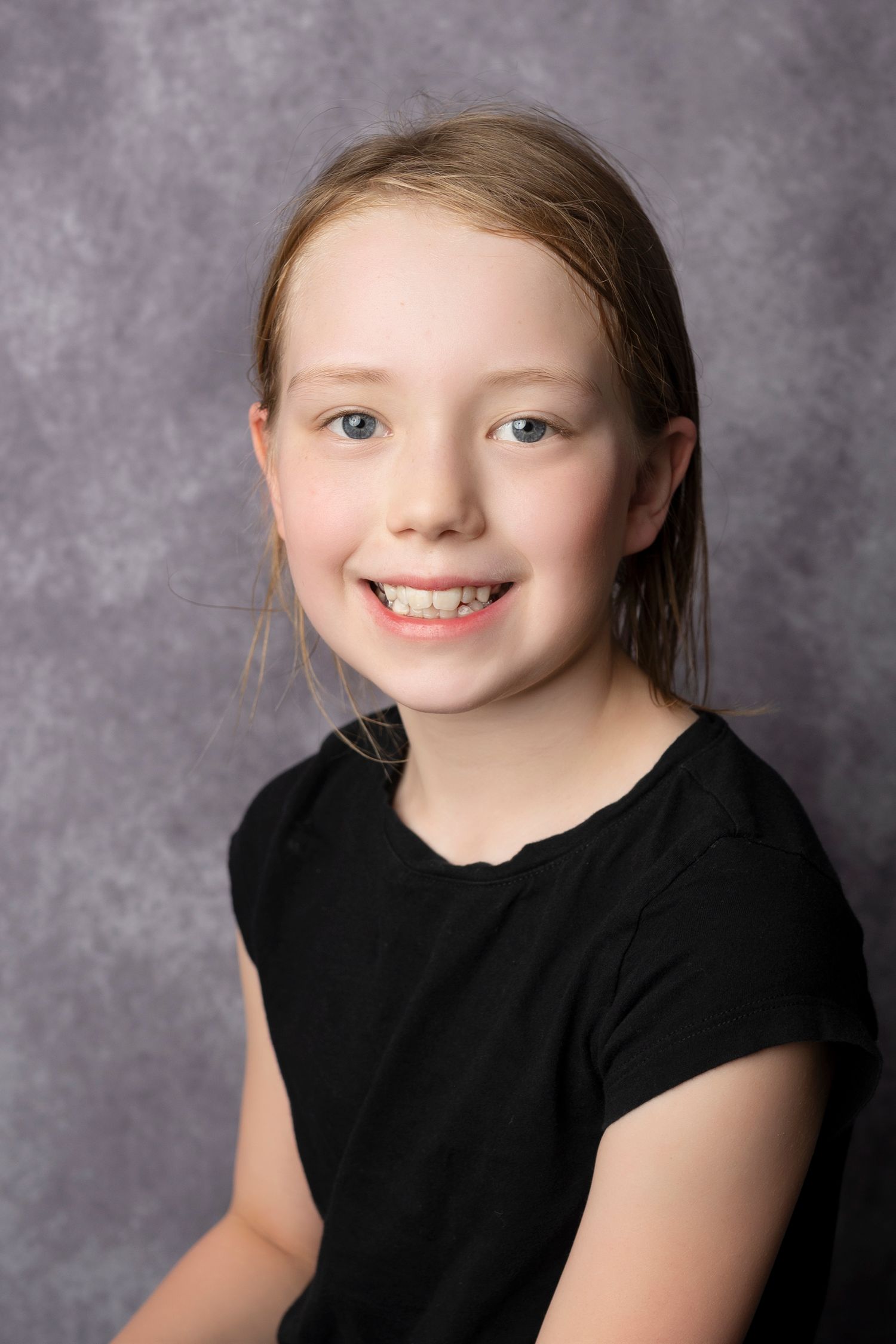 Smiling young girl with blonde hair wearing a black shirt, photographed against a gray studio background.