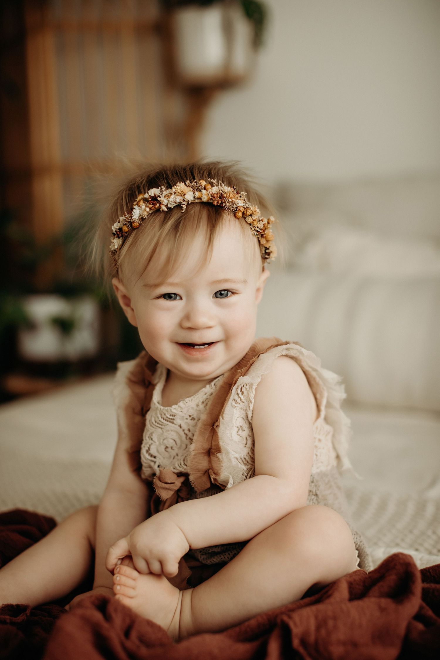Baby wearing a delicate floral crown and lace dress smiles while laying on white bedding.