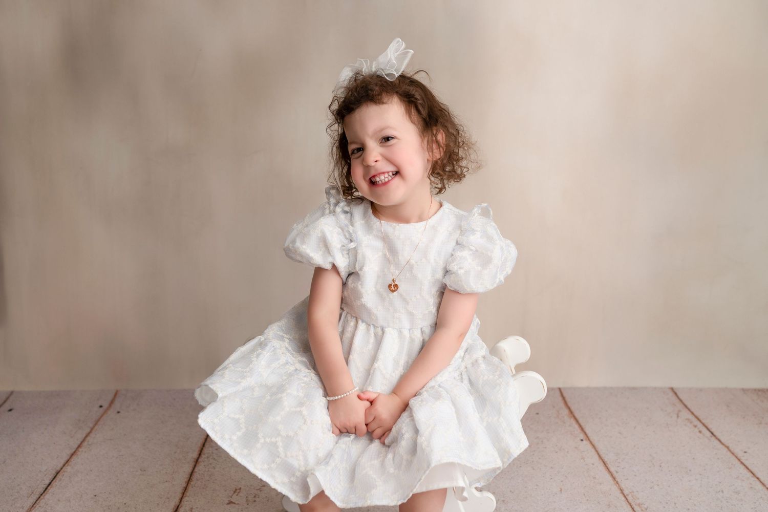 A baby in a white lace dress sits and smiles on a light wooden floor against a neutral backdrop.
