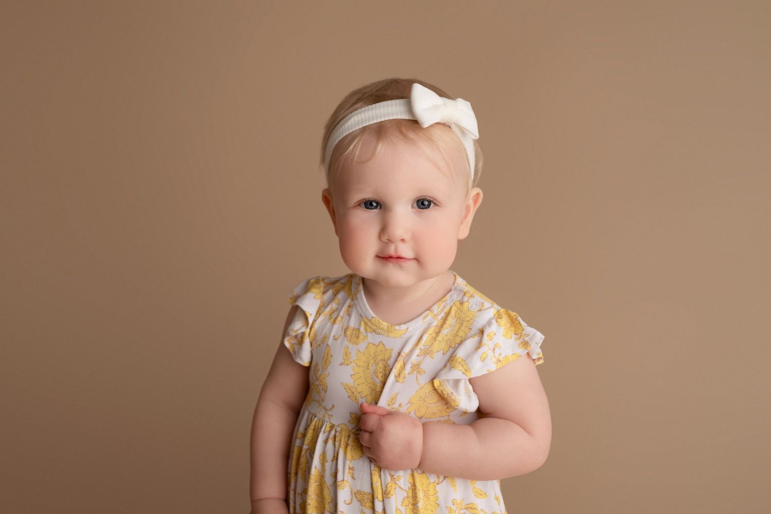 Baby wearing a yellow and white polka dot dress sits on a brown backdrop in a series of candid poses.
