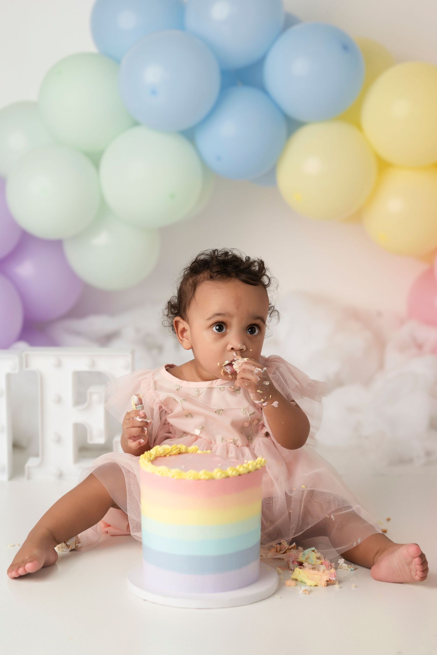 A baby in pink tulle enjoying a pastel rainbow cake with balloon arch decoration in soft colors.
