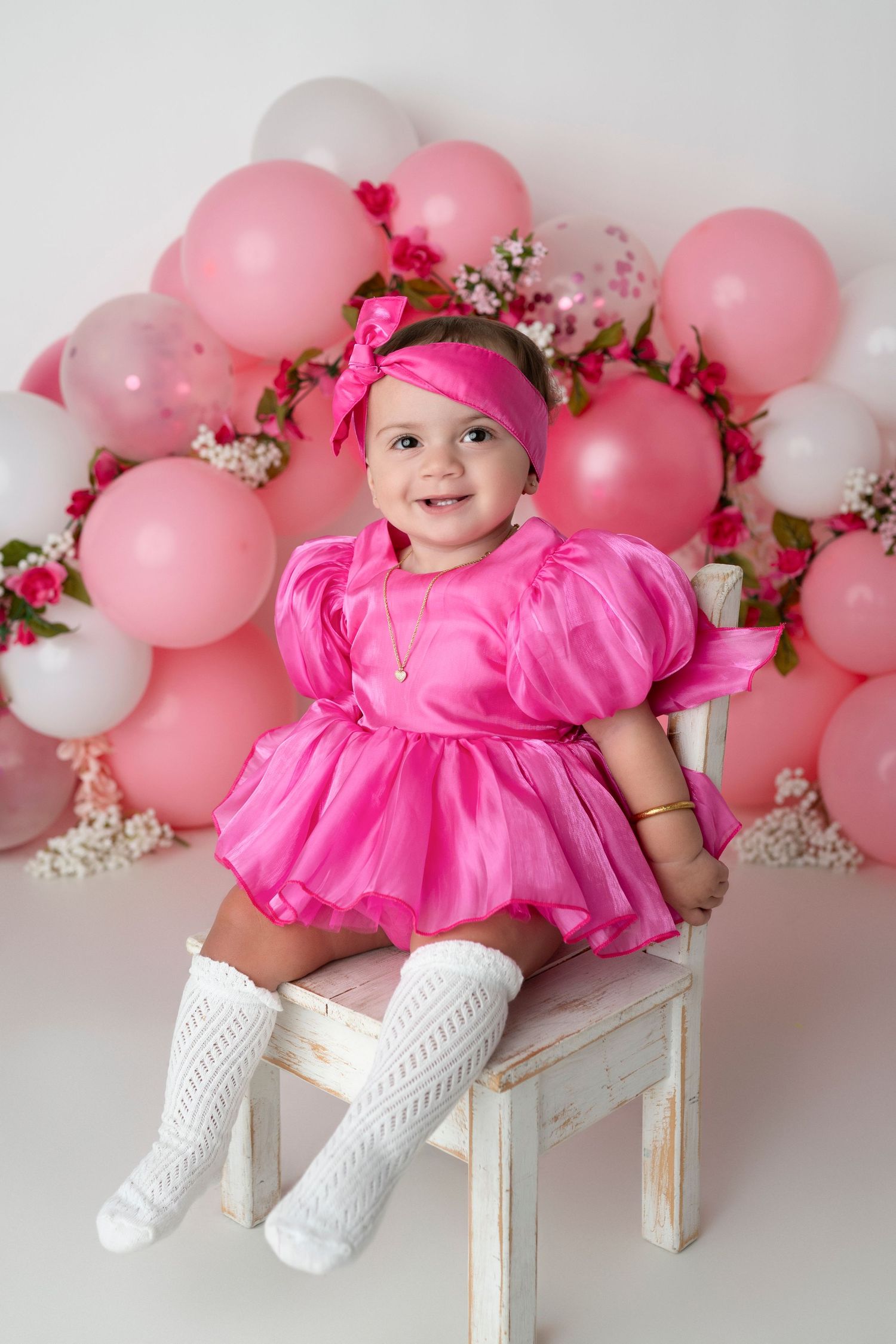 Baby in pink dress poses on white bench with pink and white balloon garland backdrop and floral decorations.