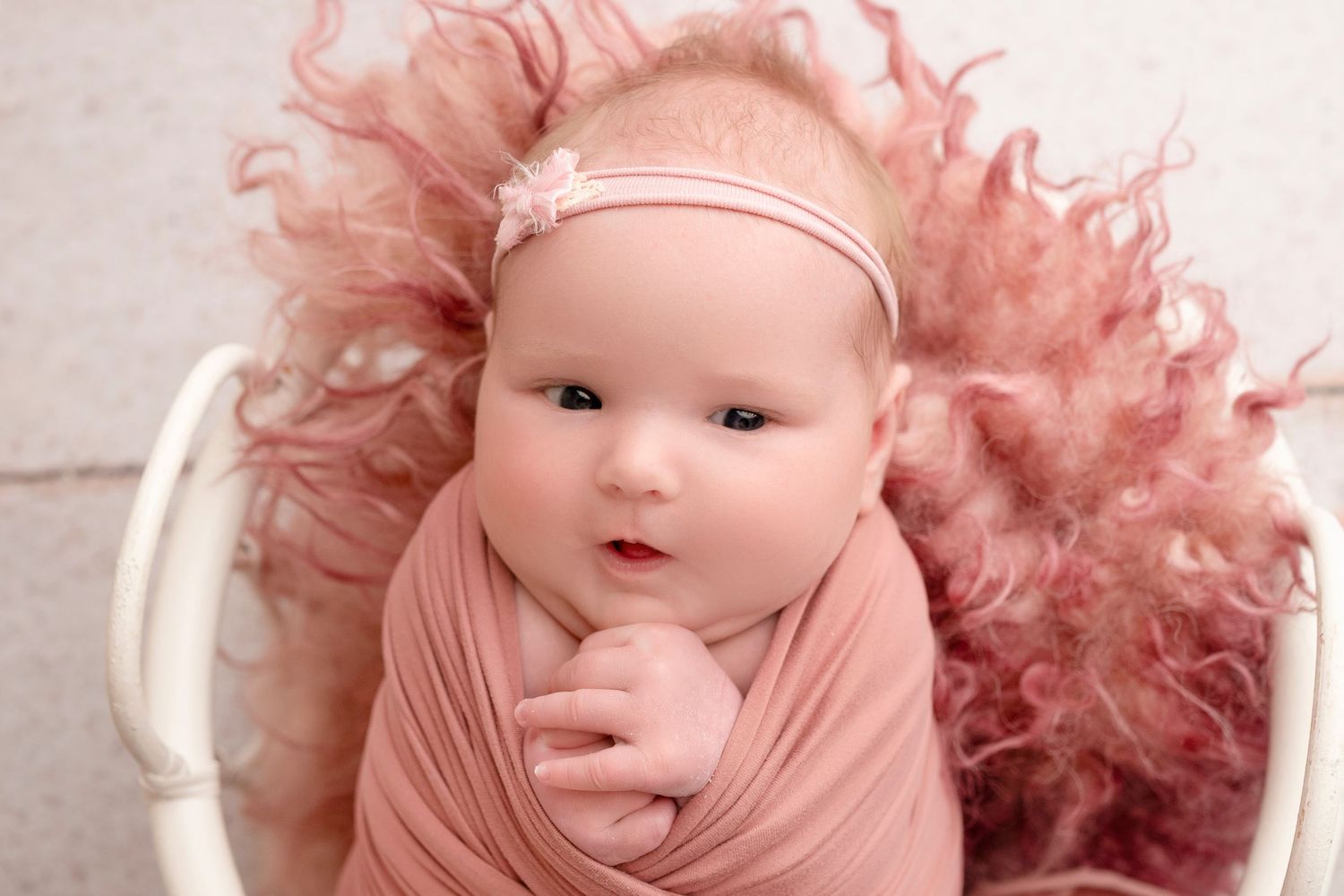 Newborn baby swaddled in pink wrap with floral headband, nestled in white basket with fluffy pink fur.