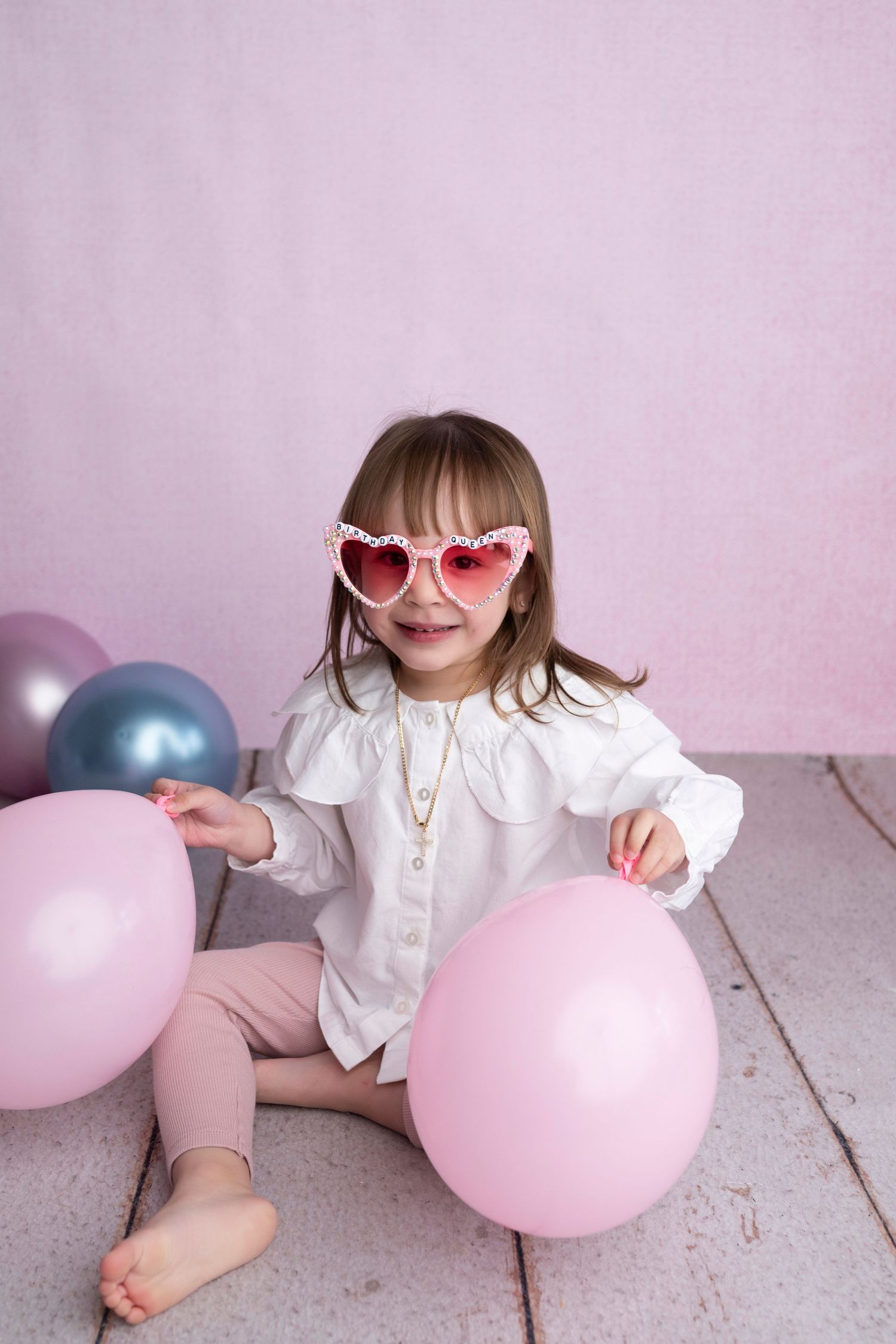 Young girl in heart-shaped sunglasses sits on floor with pink and purple balloons against pink background.