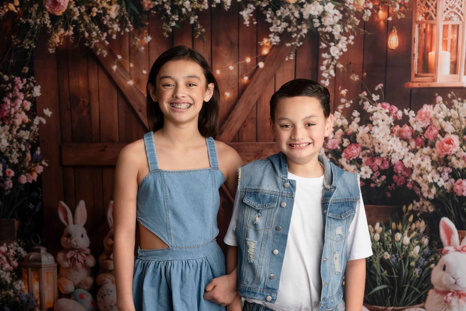 Two smiling children pose in denim outfits before a floral barn backdrop with Easter bunnies.