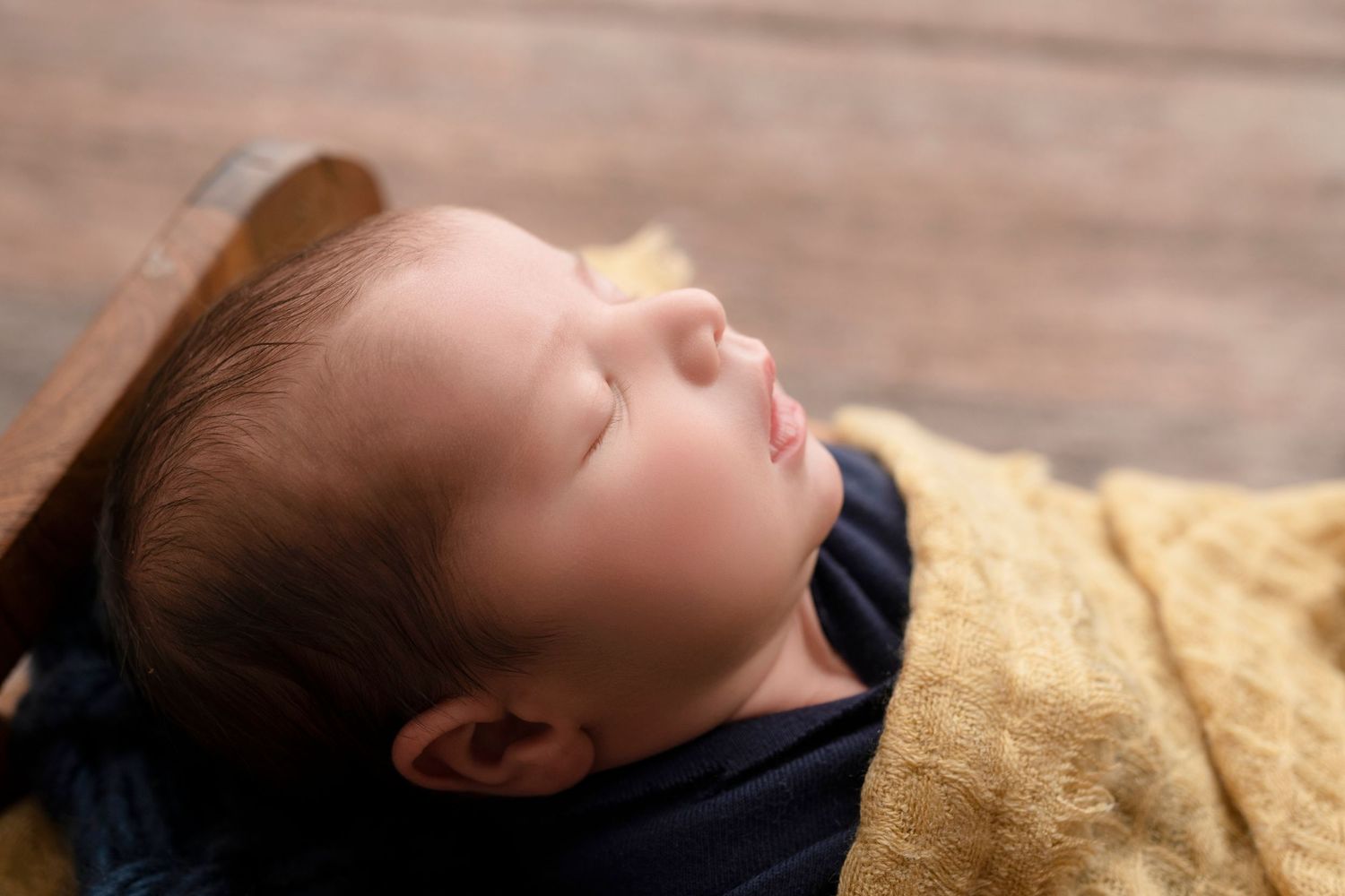 Peaceful newborn baby sleeping wrapped in a golden yellow blanket, resting in a wooden bowl prop.