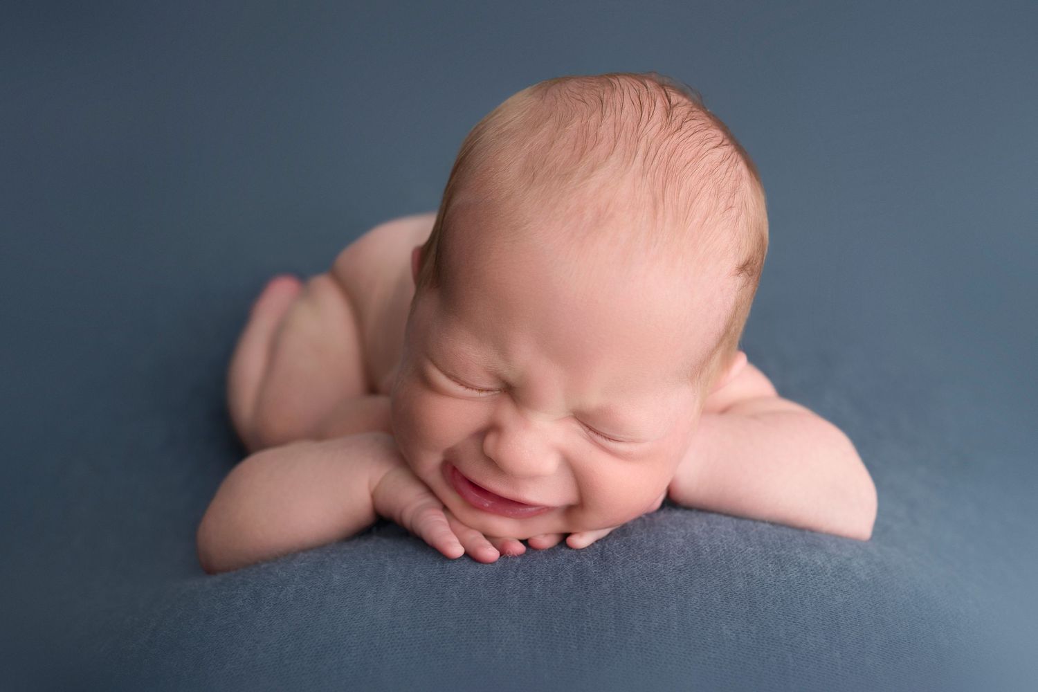 Smiling newborn baby lying on blue fabric with chin resting on hands in a peaceful posed portrait.