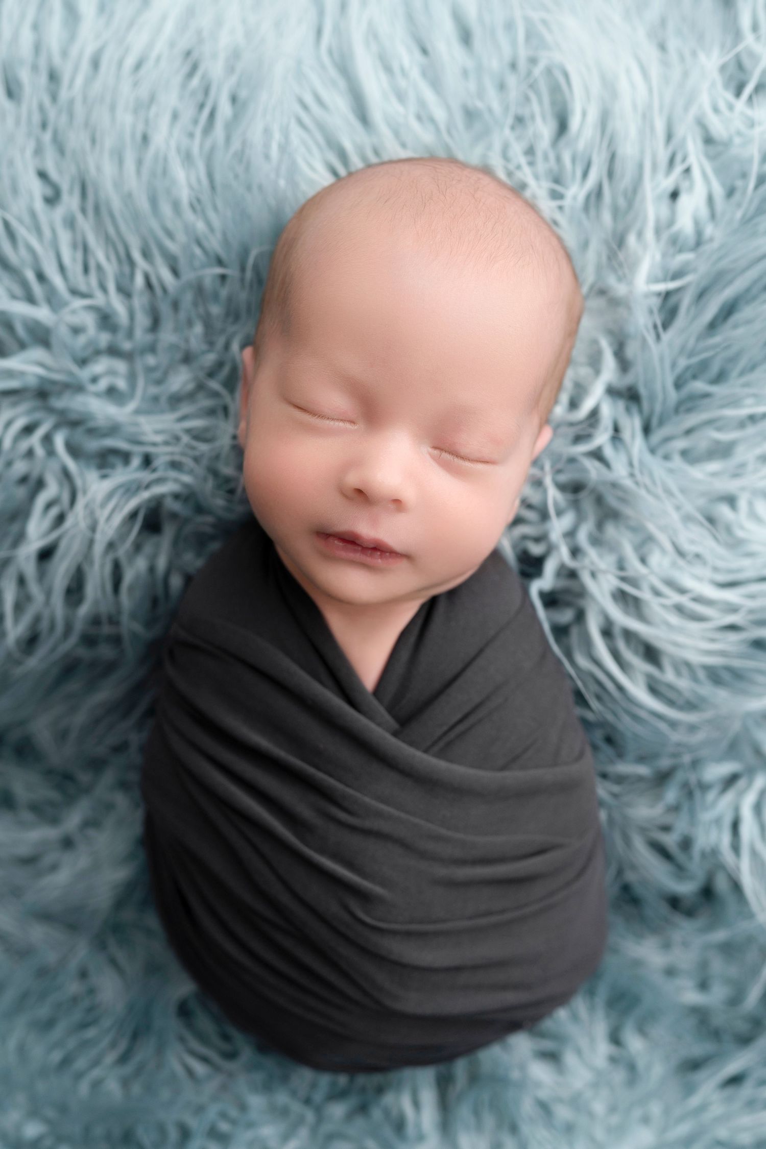 Newborn baby swaddled in gray wrap sleeping peacefully on a soft blue fluffy rug in a professional photo.