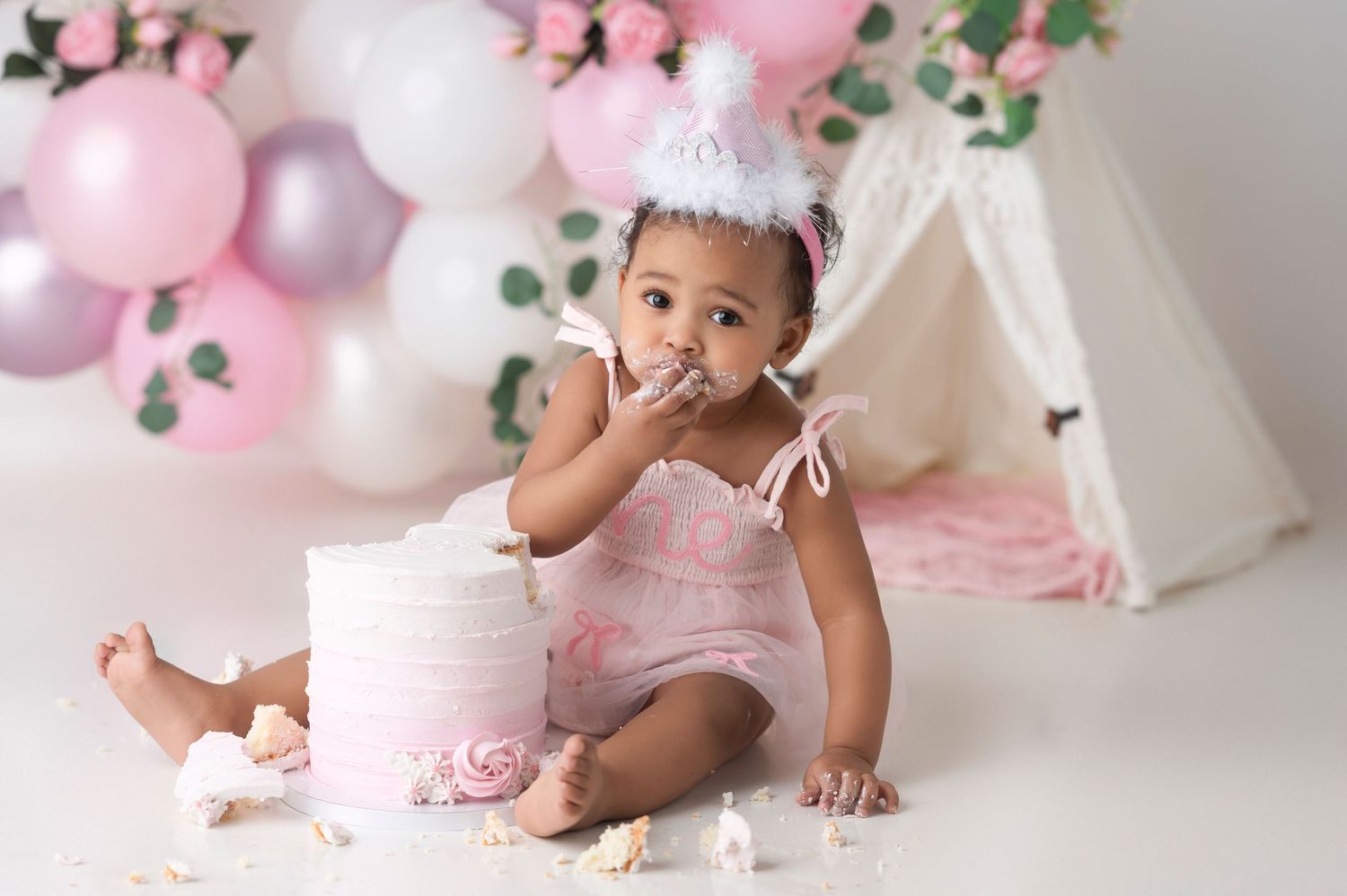 Baby girl in pink romper eating first birthday smash cake surrounded by pink balloons and teepee backdrop.