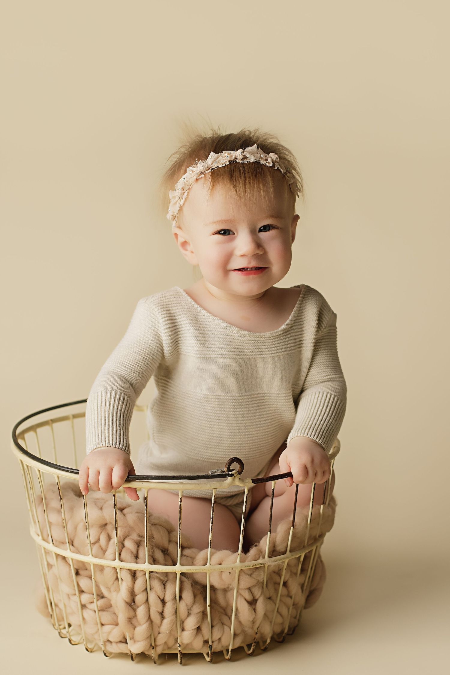 A small child wearing a beige sweater sits in a woven basket against a neutral background, wearing a delicate floral headband.
