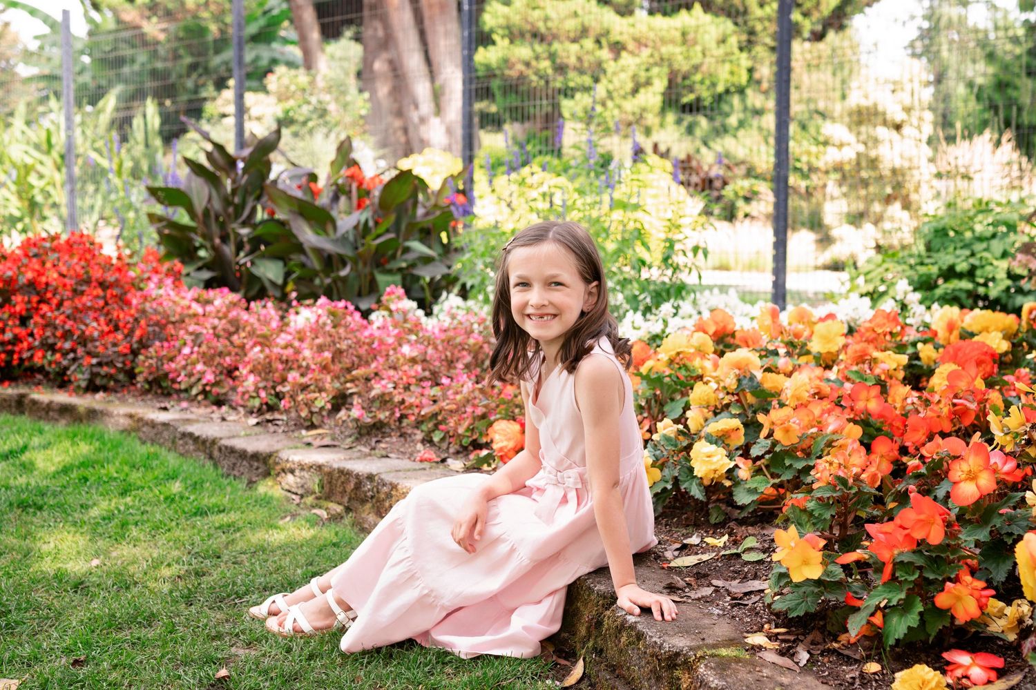 A girl in a pink dress sits among vibrant orange and red flowers in a lush garden setting.