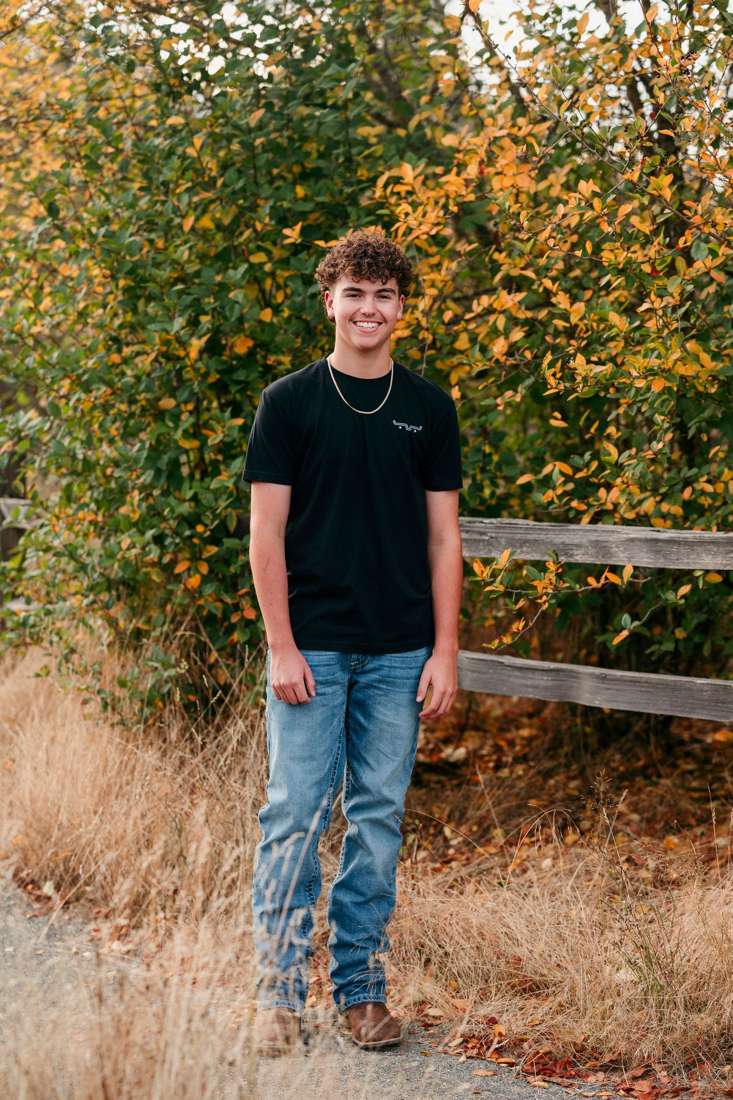 Person in casual attire standing by autumn foliage and wooden fence in natural outdoor setting.