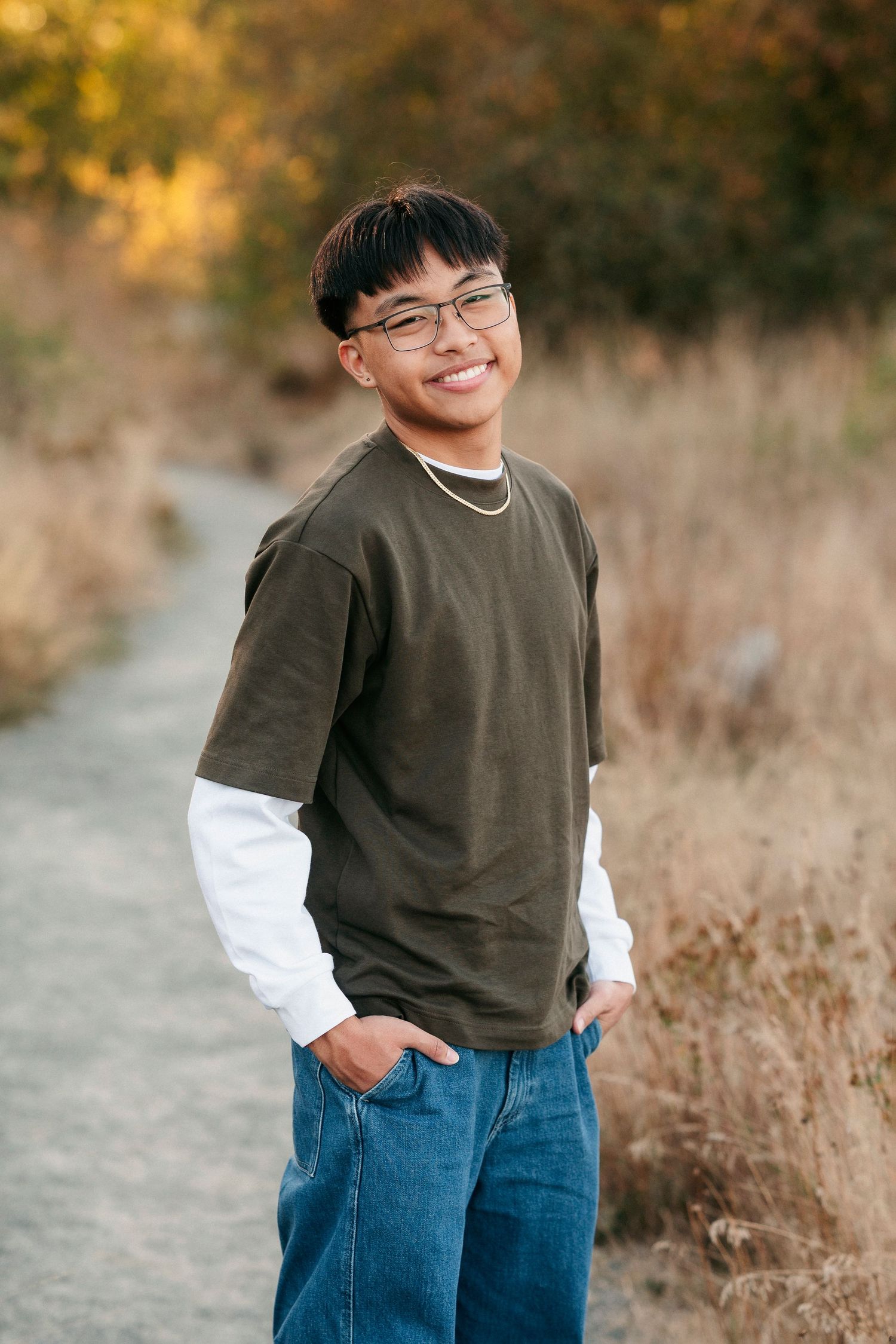 A person wearing a brown sweater and jeans poses outdoors in autumn against a blurred natural background.