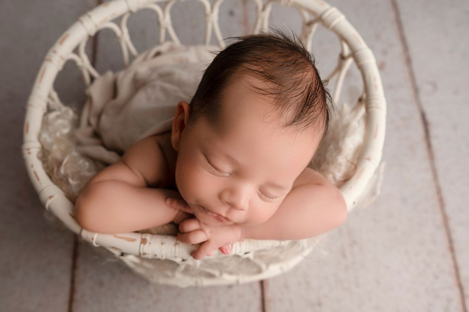 A sleeping newborn baby lies curled up in a cozy cream-colored basket on a wooden floor.