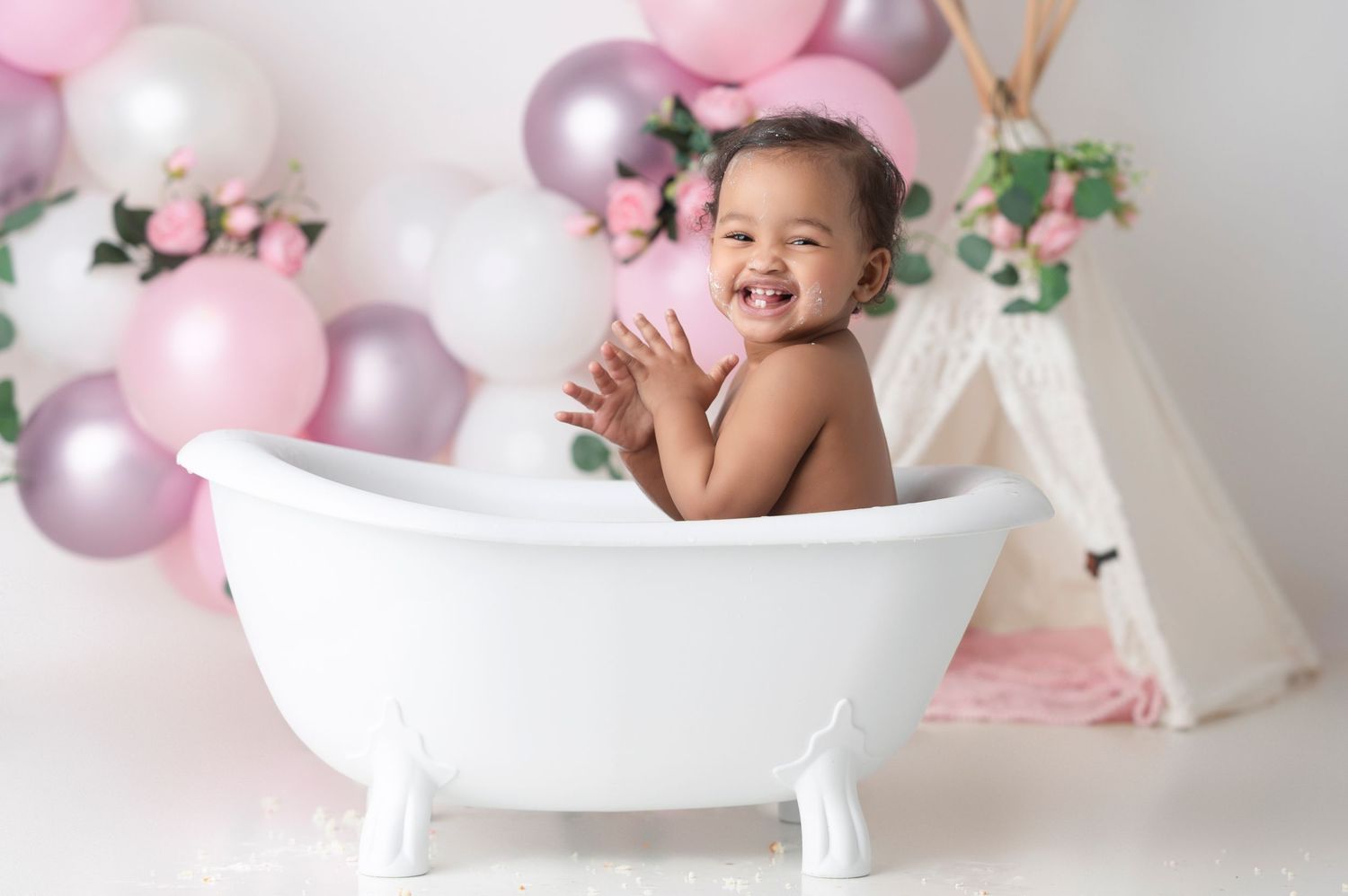 Smiling toddler sitting in a white clawfoot bathtub surrounded by pink and purple balloons.