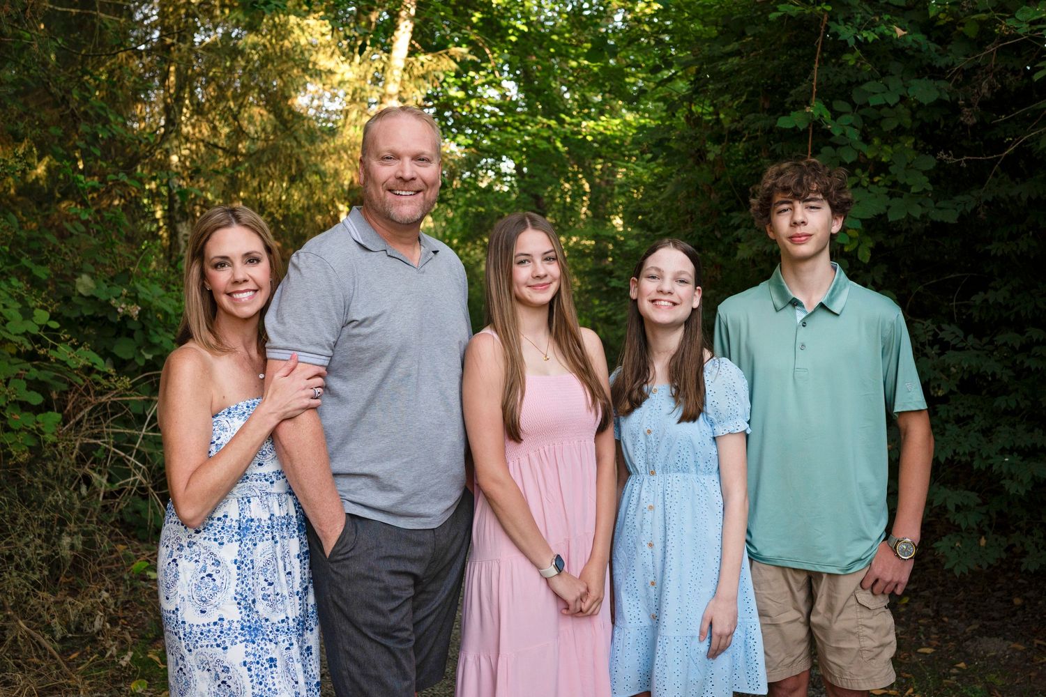 A family wearing coordinated pastel outfits poses together outdoors in a lush green forest setting.
