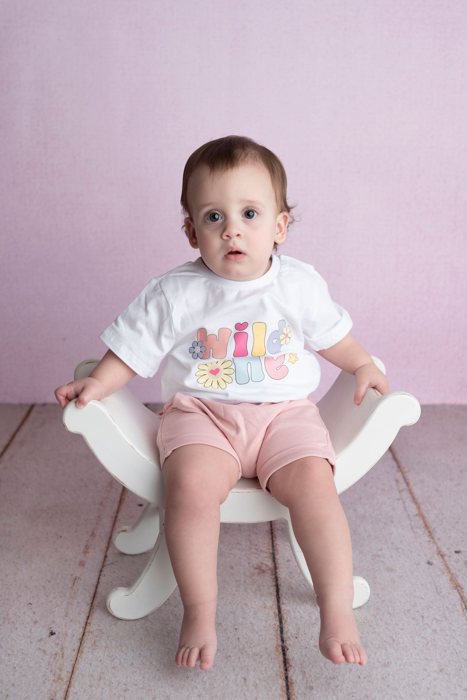 Baby girl in white graphic tee and pink shorts sits in white rocker chair against pink background.