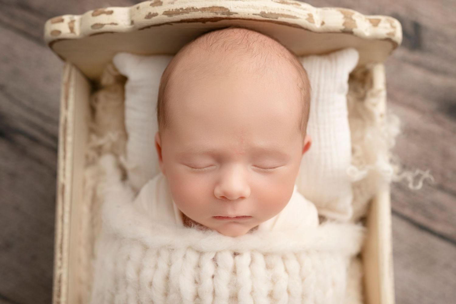 Peaceful newborn baby sleeping in a tiny cream wooden bed wrapped in a soft white knit blanket.
