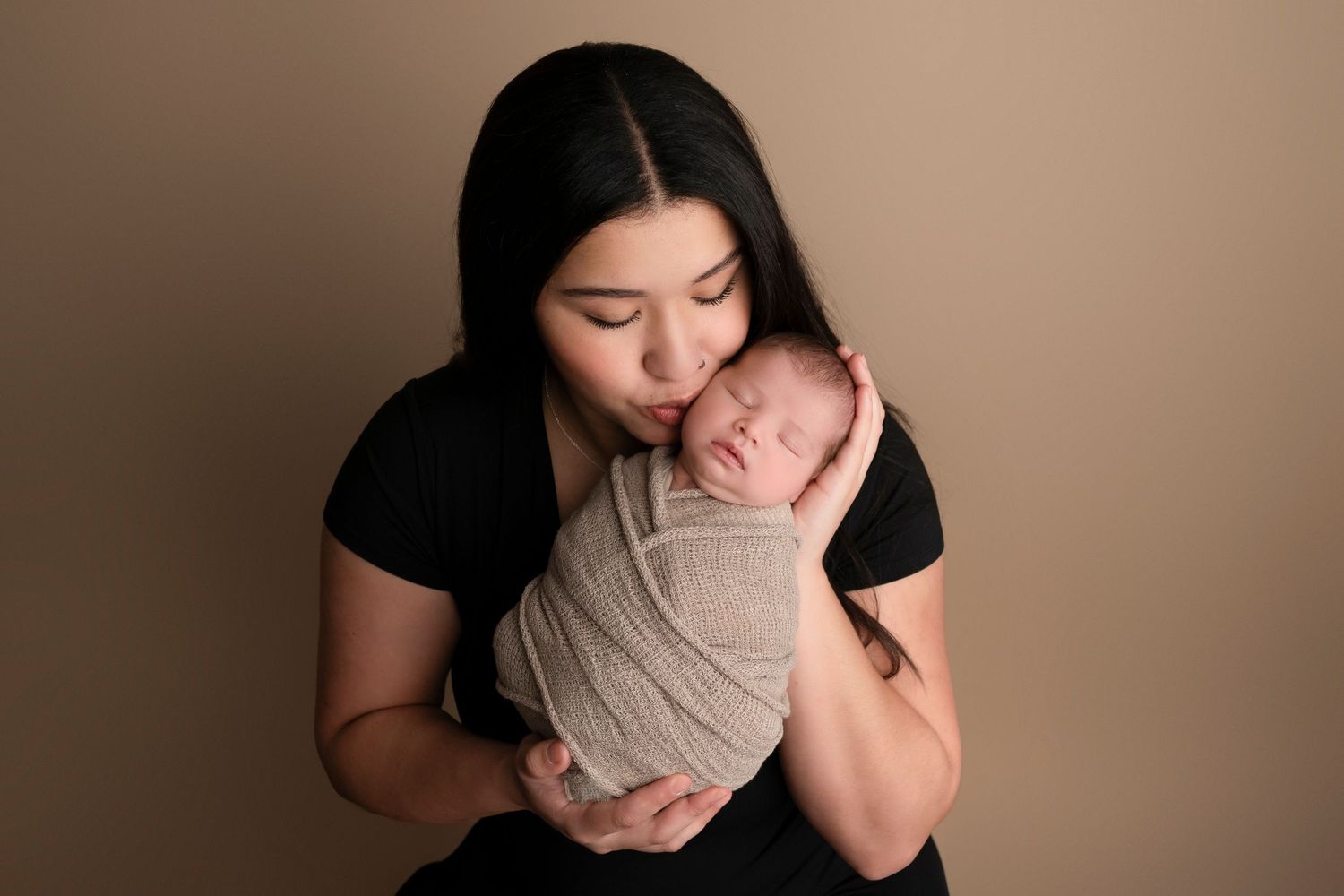 A mother in a black dress tenderly kisses her swaddled newborn baby against a warm beige studio background.