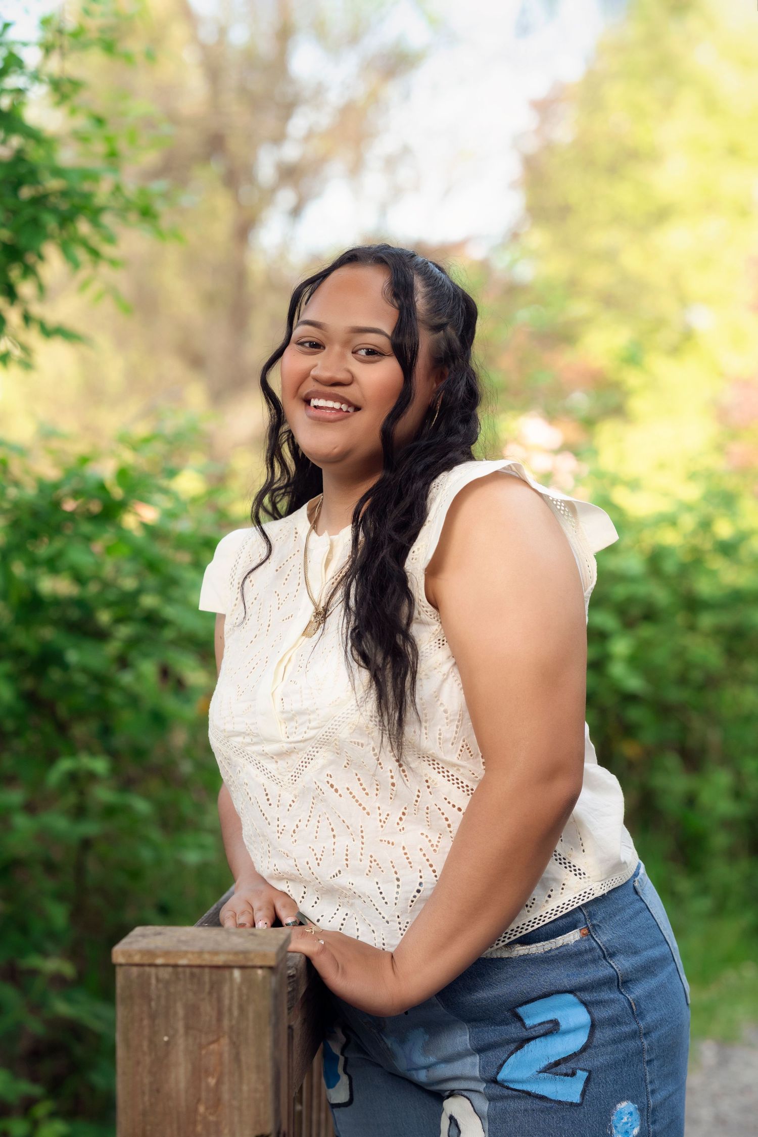 A joyful person in a white lace top and denim poses outdoors against a blurred natural background.
