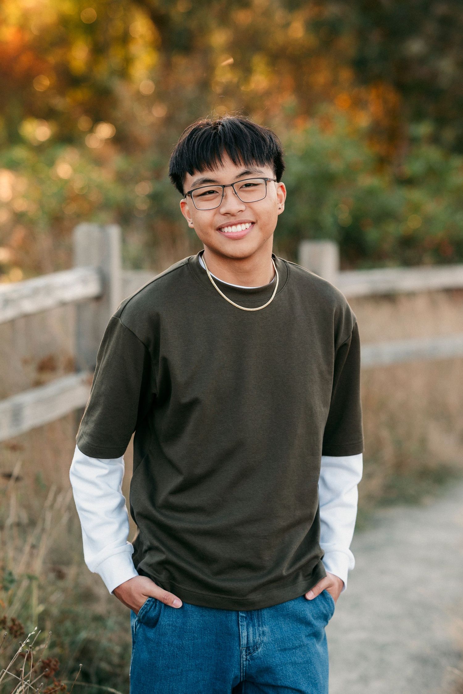 Person wearing glasses, black sweater and jeans poses outdoors during autumn with warm bokeh lighting in background.