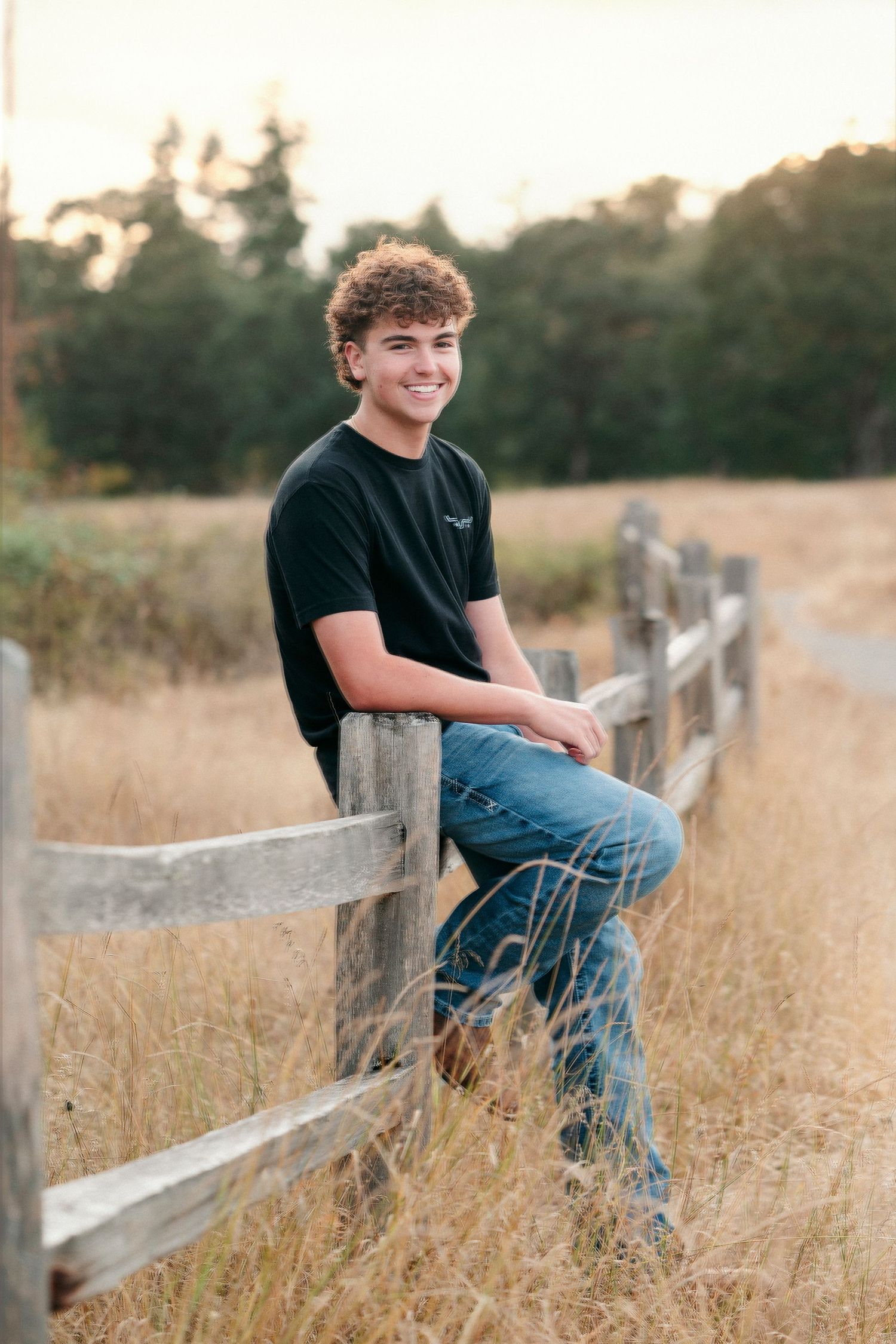 A rustic outdoor portrait shows someone sitting on a wooden fence in a golden field at sunset.