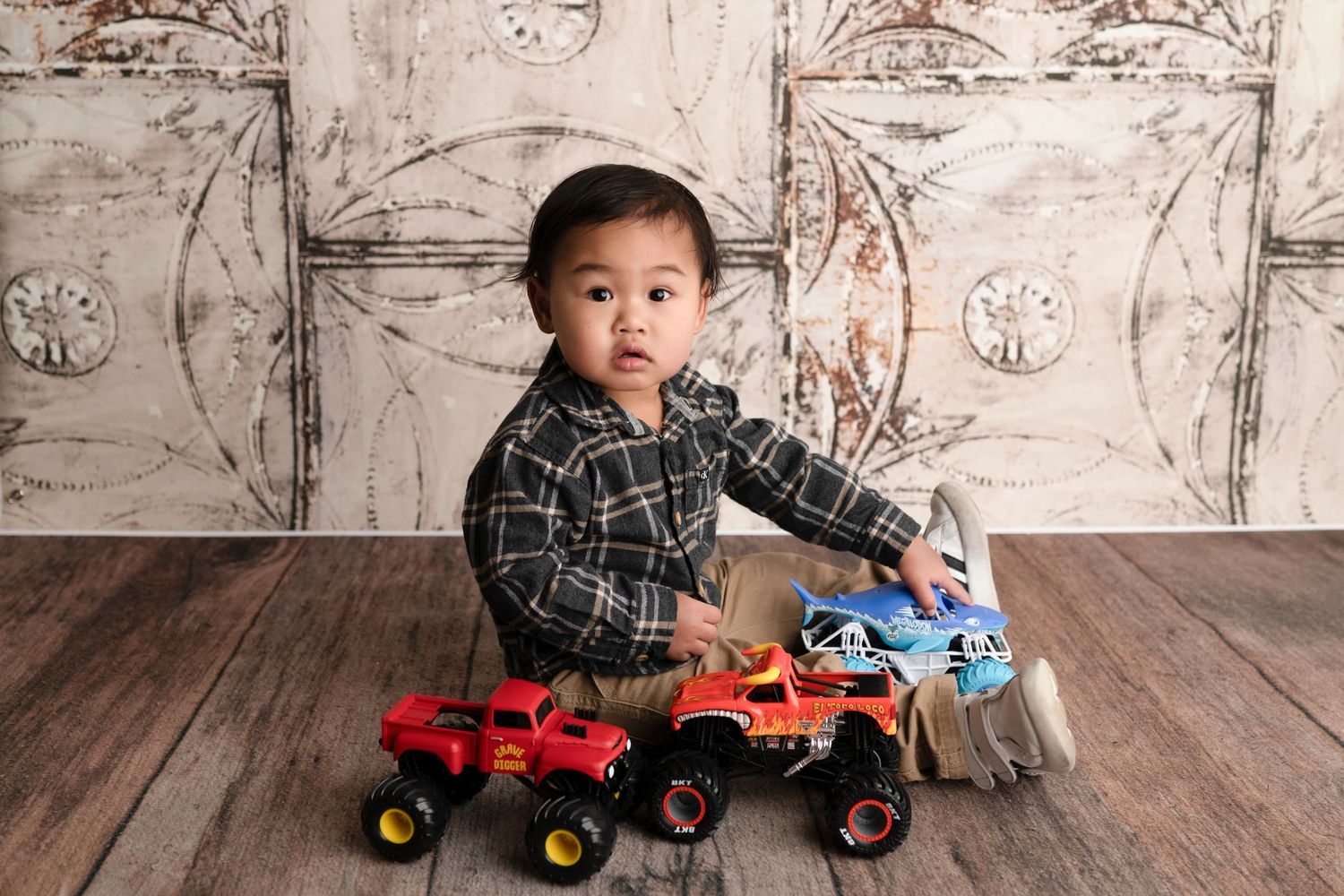 Toddler boy in plaid shirt playing with colorful monster truck toys on wood floor against rustic white backdrop.