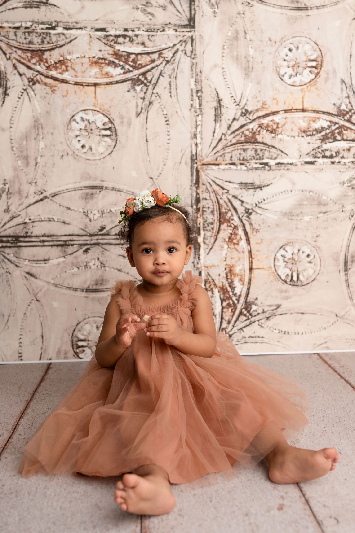 A toddler girl in a dusty rose tulle dress and floral headband sits barefoot against a vintage decorative backdrop.