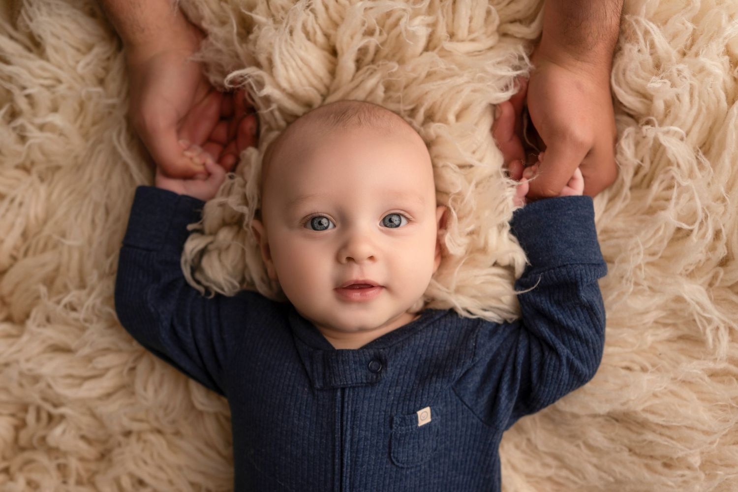 Baby in navy blue sweater lying on soft beige textured rug while playing and smiling at camera.